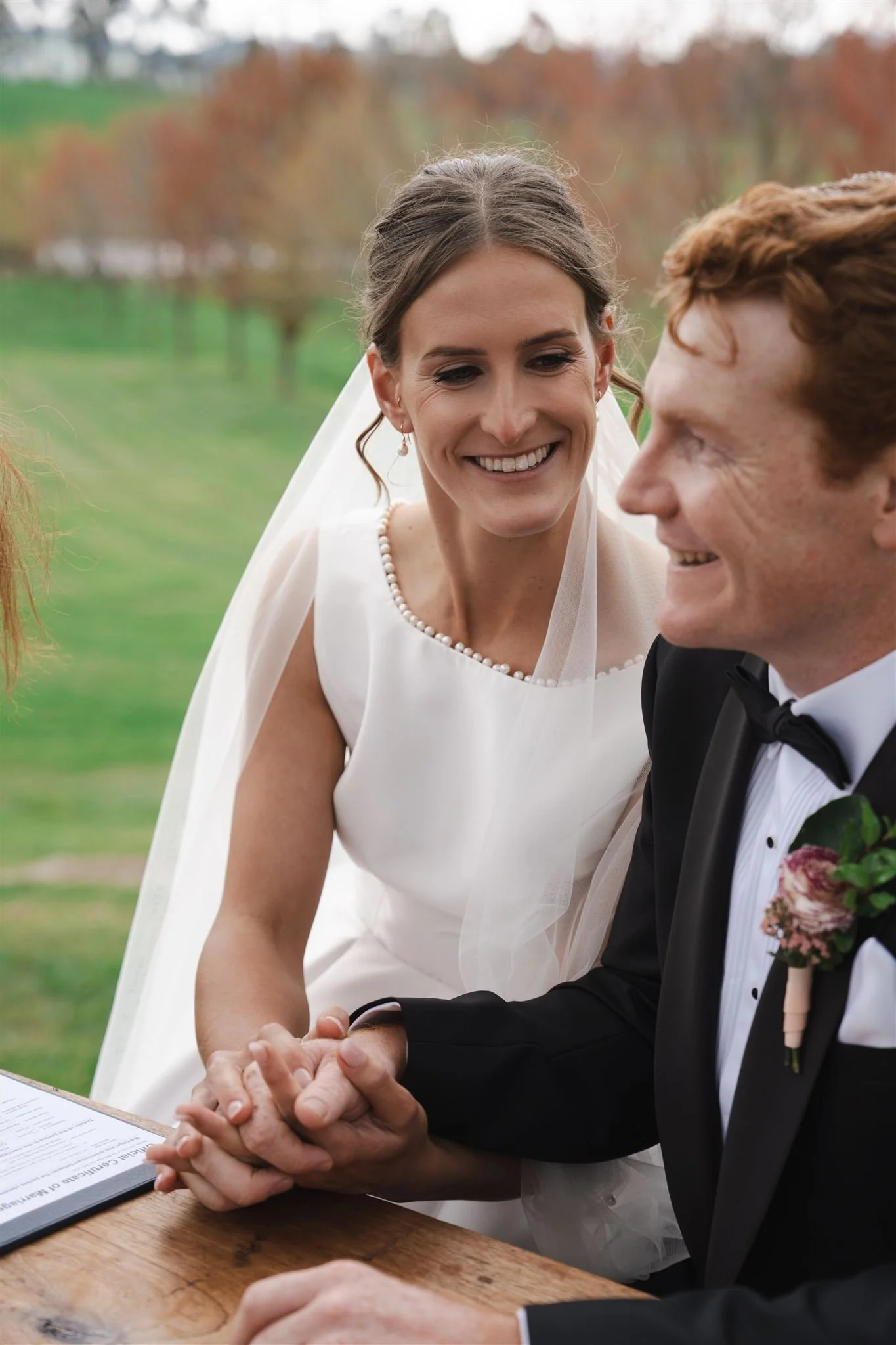 A bride and groom holding hands outdoors during their wedding ceremony, smiling at each other with a scenic landscape in the background, vows, ceremony, wedding signing.