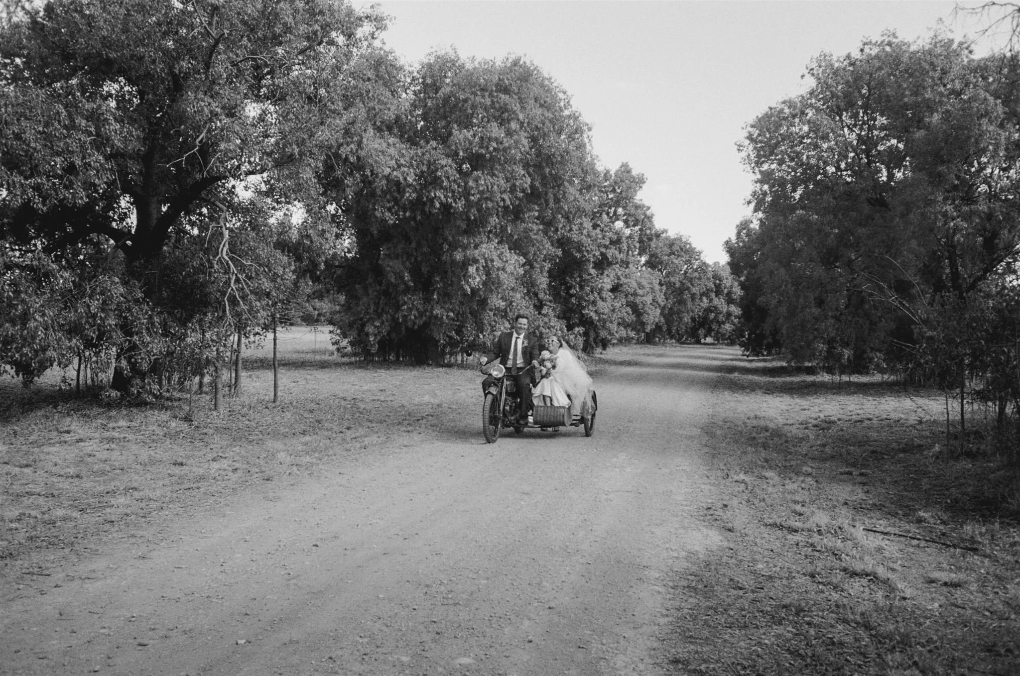 Bride and groom, country wedding, Australia, private property, Black and white photo of a man in a suit riding a bicycle with a sidecar, carrying a bride in a wedding dress through a wooded area with large trees and a dirt path.