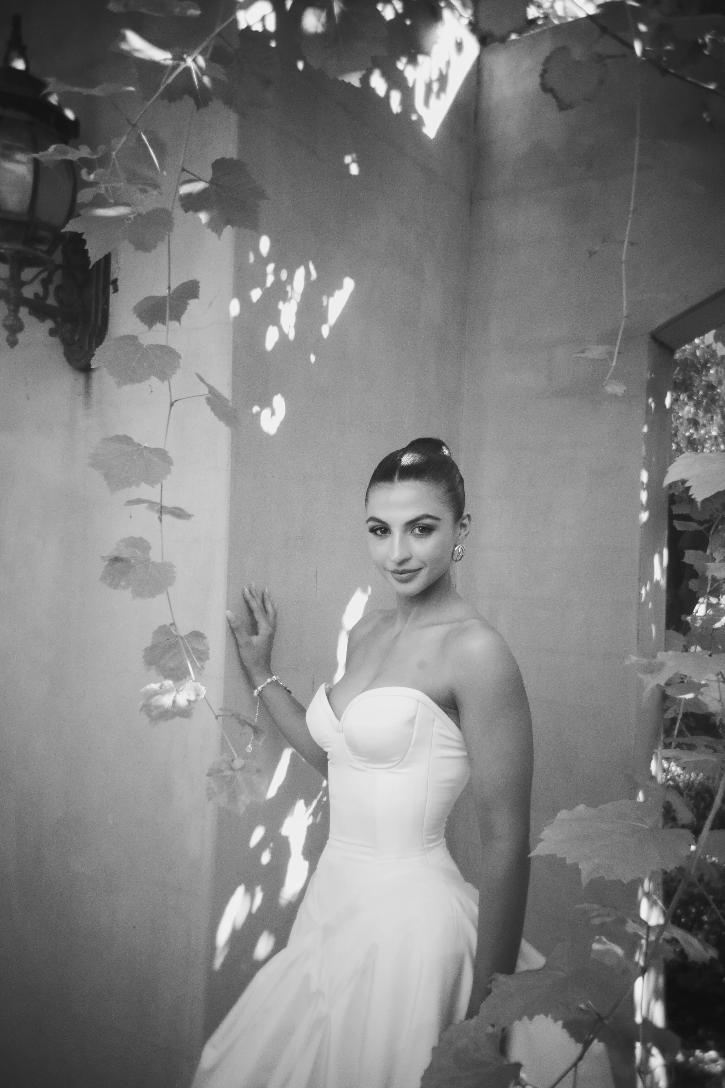 A woman in a strapless wedding dress standing by a wall with leaves casting shadows, looking at the camera with a slight smile.