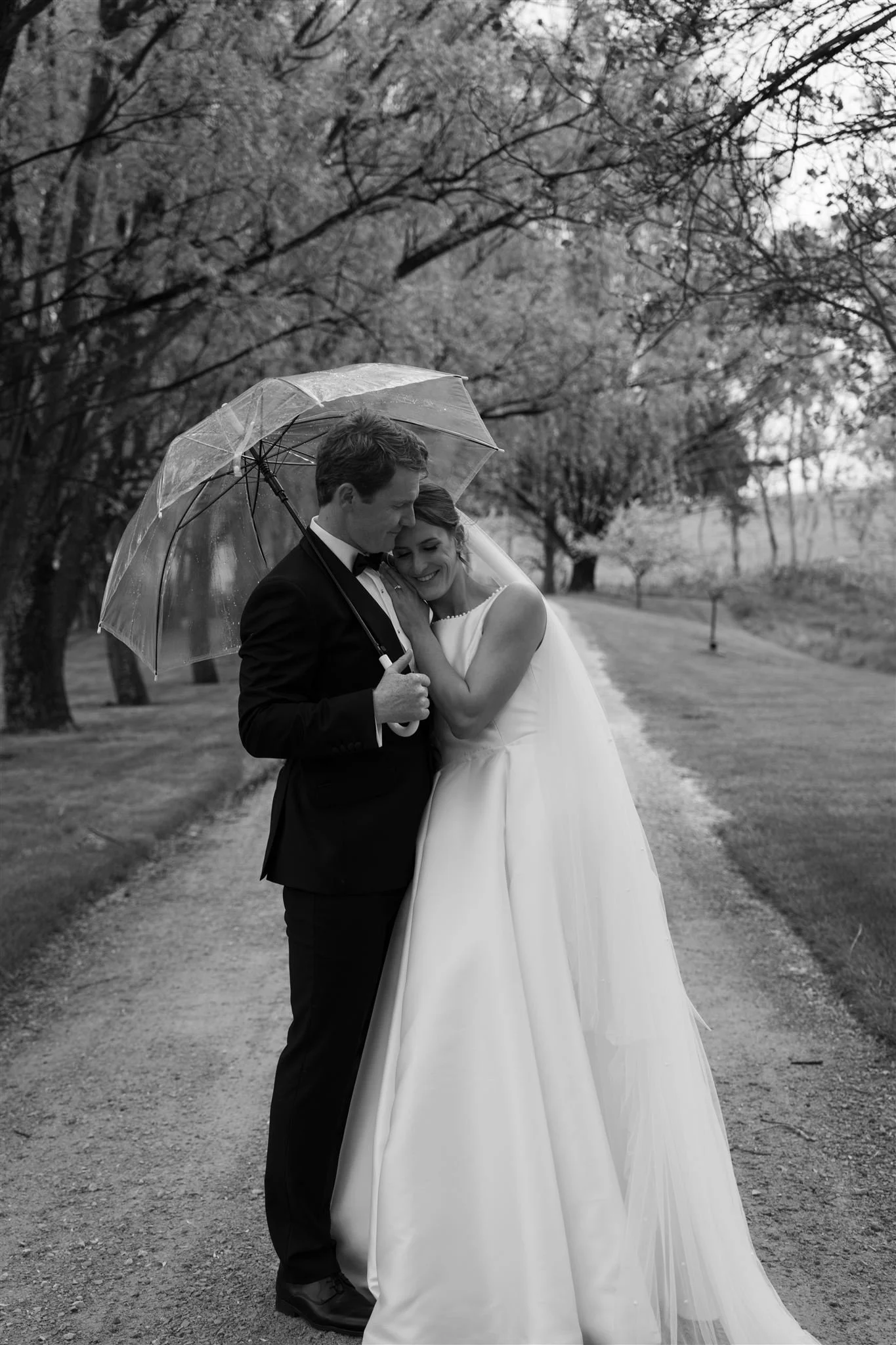 couple portraits, A black and white photograph of a bride and groom standing close together under a transparent umbrella on a dirt path in a park with trees and grass in the background, taking a tender moment on their wedding day.