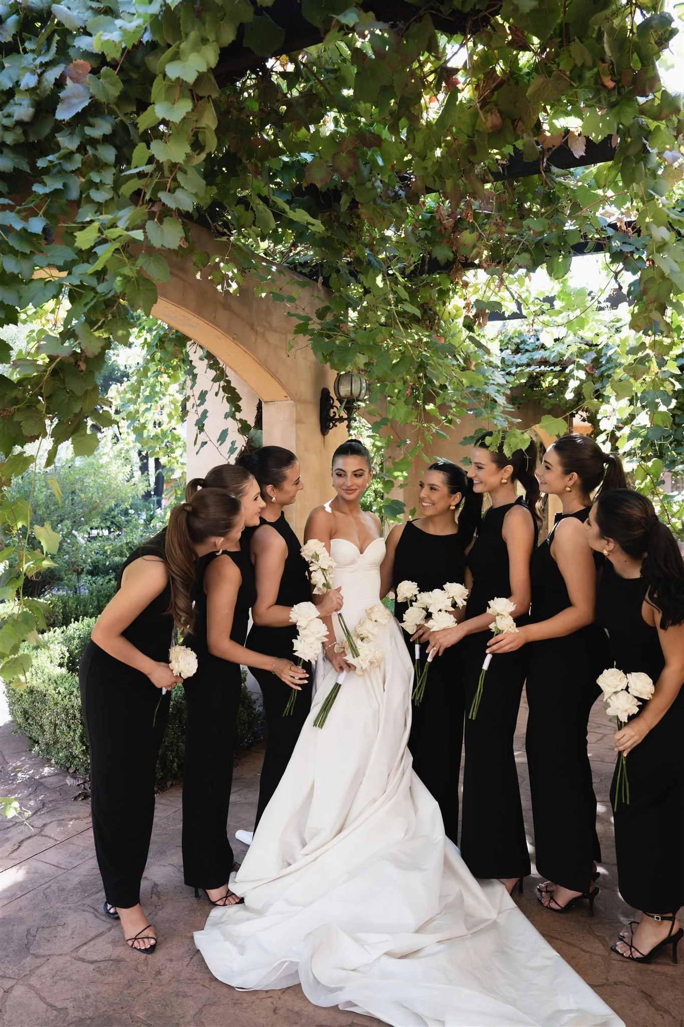 Bridal party, Bride in a white wedding gown and six bridesmaids in black dresses holding white flowers, standing under green leafy vines in a garden setting.