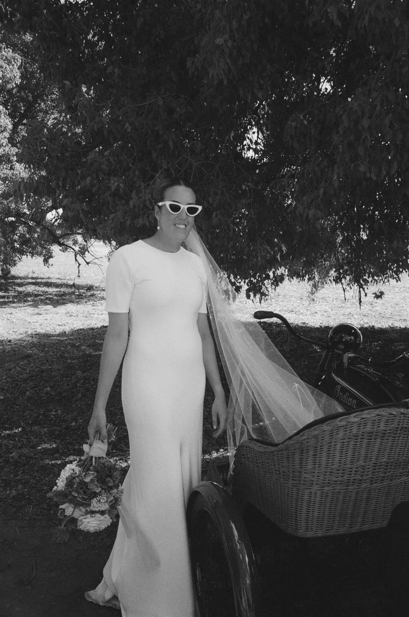 A woman in a white wedding dress with sunglasses and earrings, holding a bouquet, standing beside a rickshaw with a motorcycle in a park shaded by trees.