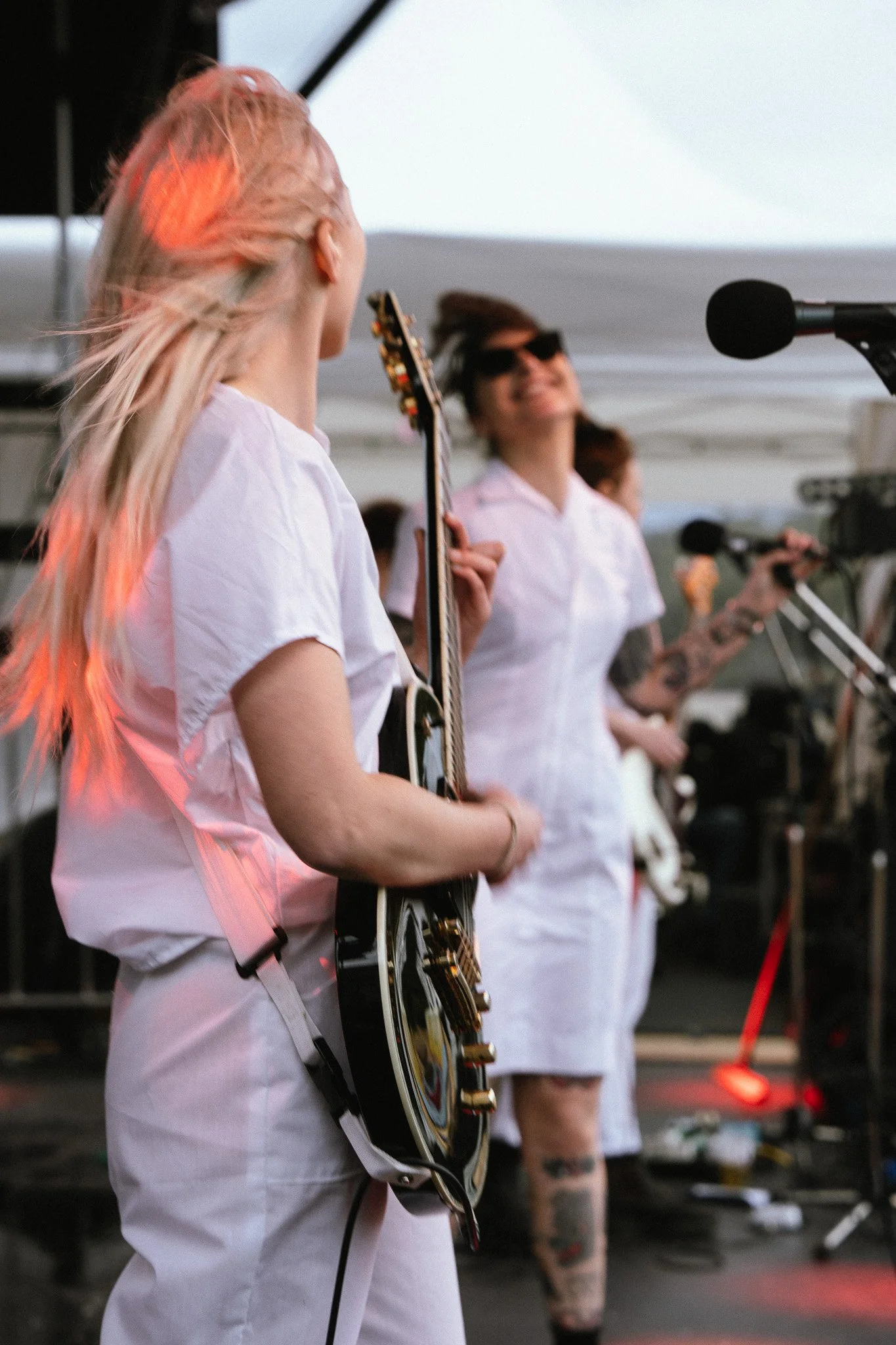 Festival content, photography. Musicians performing on stage, wearing white outfits, with guitars and microphones, under a tent.