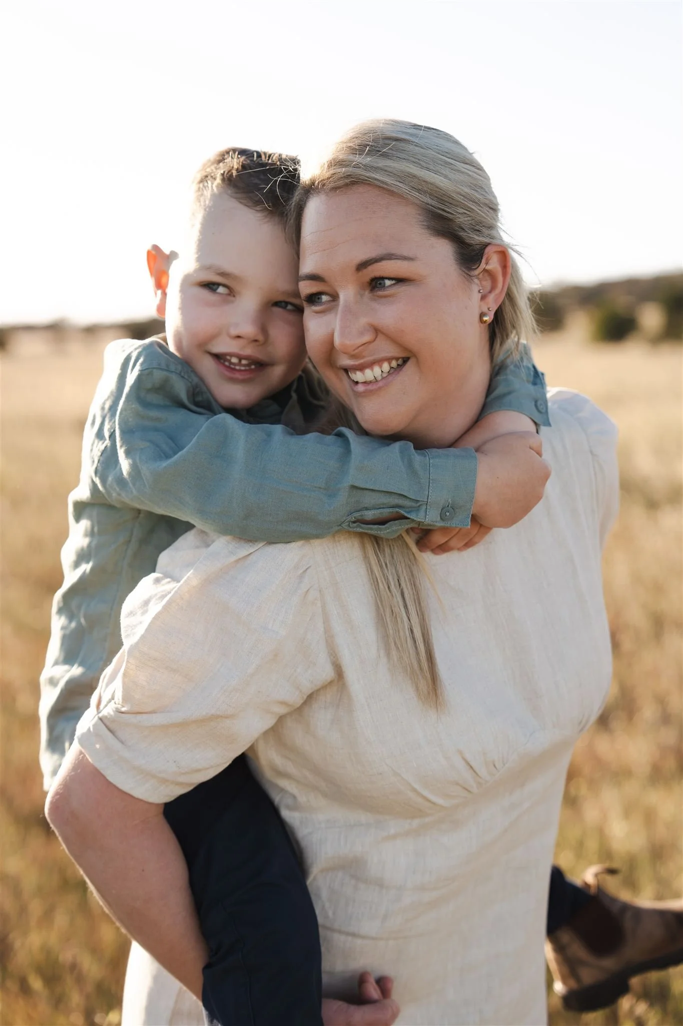 A woman carrying a young boy on her back outdoors in a field during daytime, both smiling.