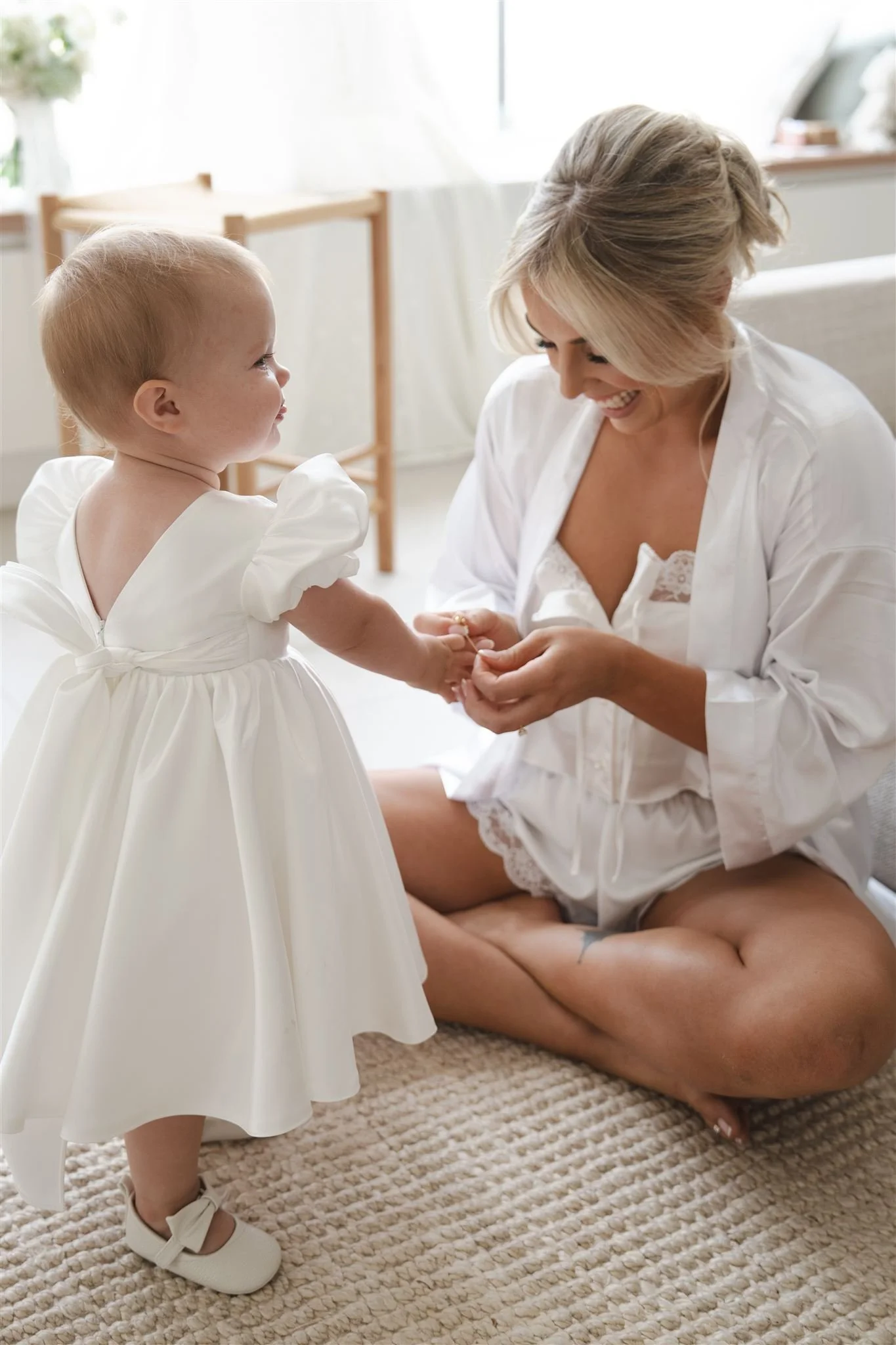 Bride and daughter, A young girl in a white dress holding hands with a smiling woman, sitting on a carpeted floor in a bright room, exchanging a ring.