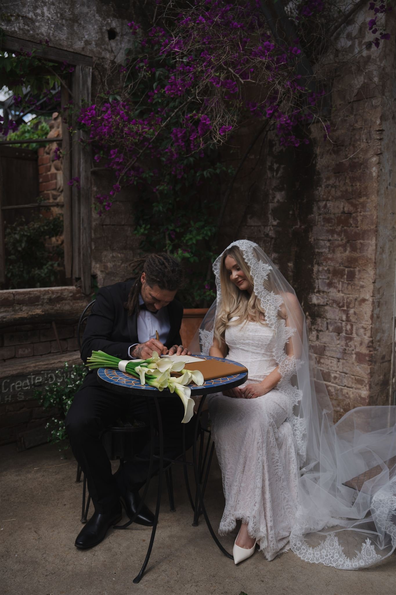 A bride in a lace wedding dress with a veil sitting at an outdoor table, signing a document, with a man in a suit sitting beside her in front of a rustic brick wall with flowering plants.