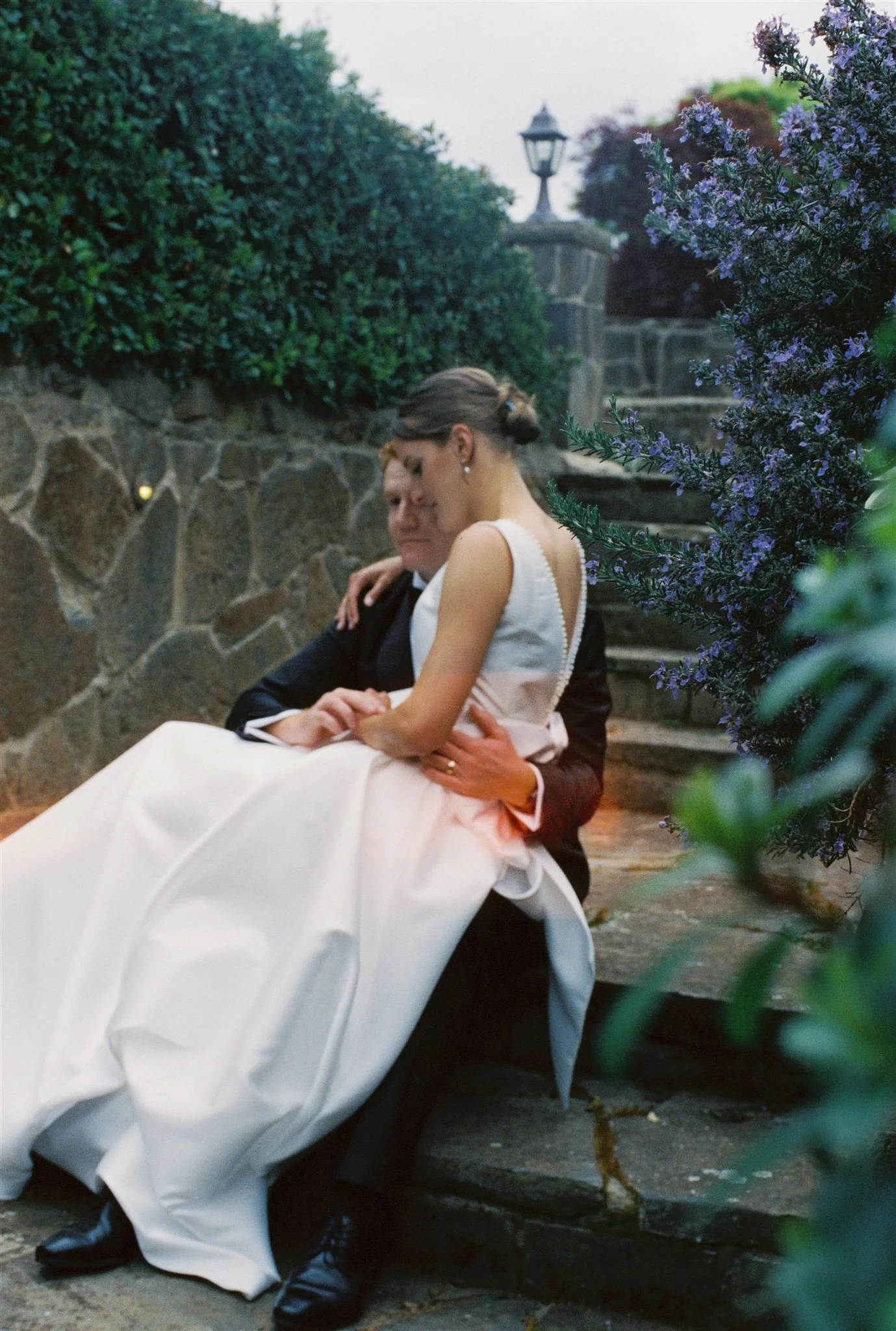 A bride and groom sitting on steps outdoors, surrounded by greenery and purple flowers, during what appears to be a wedding or romantic moment.
