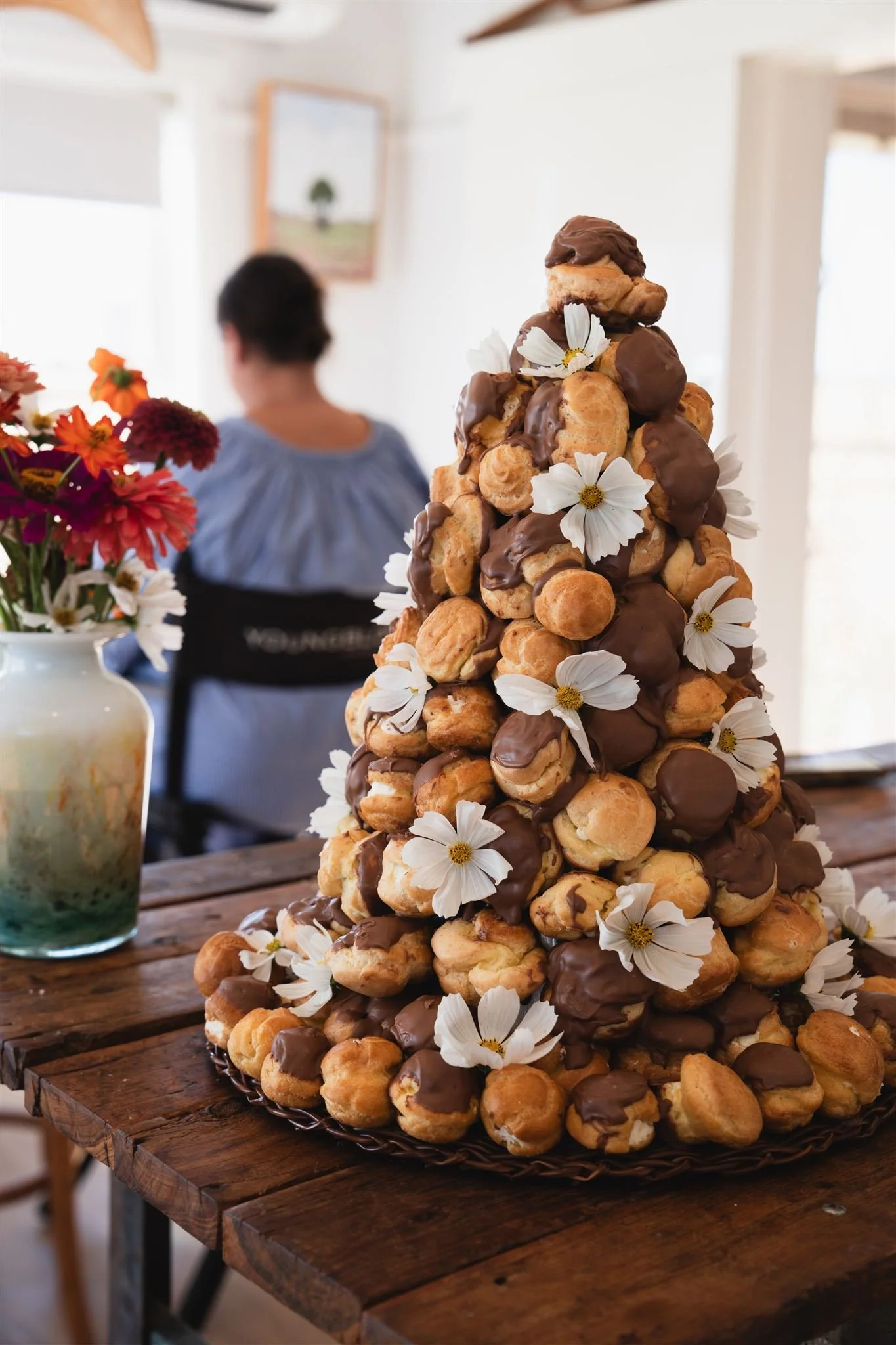 A croquembouche, wedding cake, a tower of cream puffs decorated with white flowers and drizzled with chocolate, sits on a wooden table.