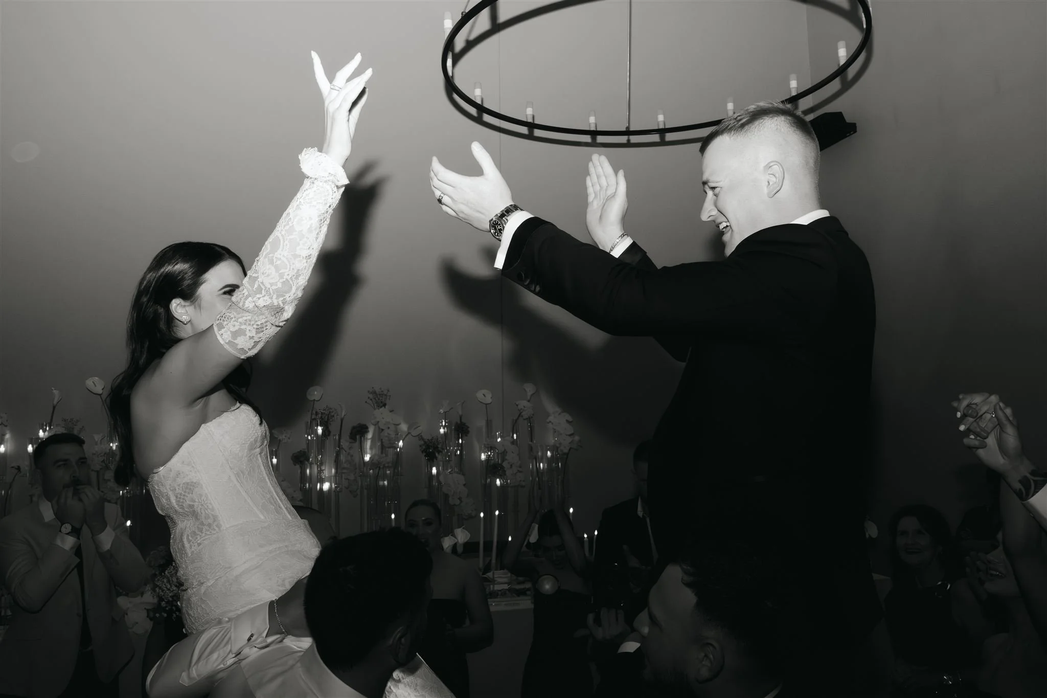 A bride and groom dancing together at their wedding reception, smiling and reaching out to each other, in a decorated indoor venue with guests watching in the background.