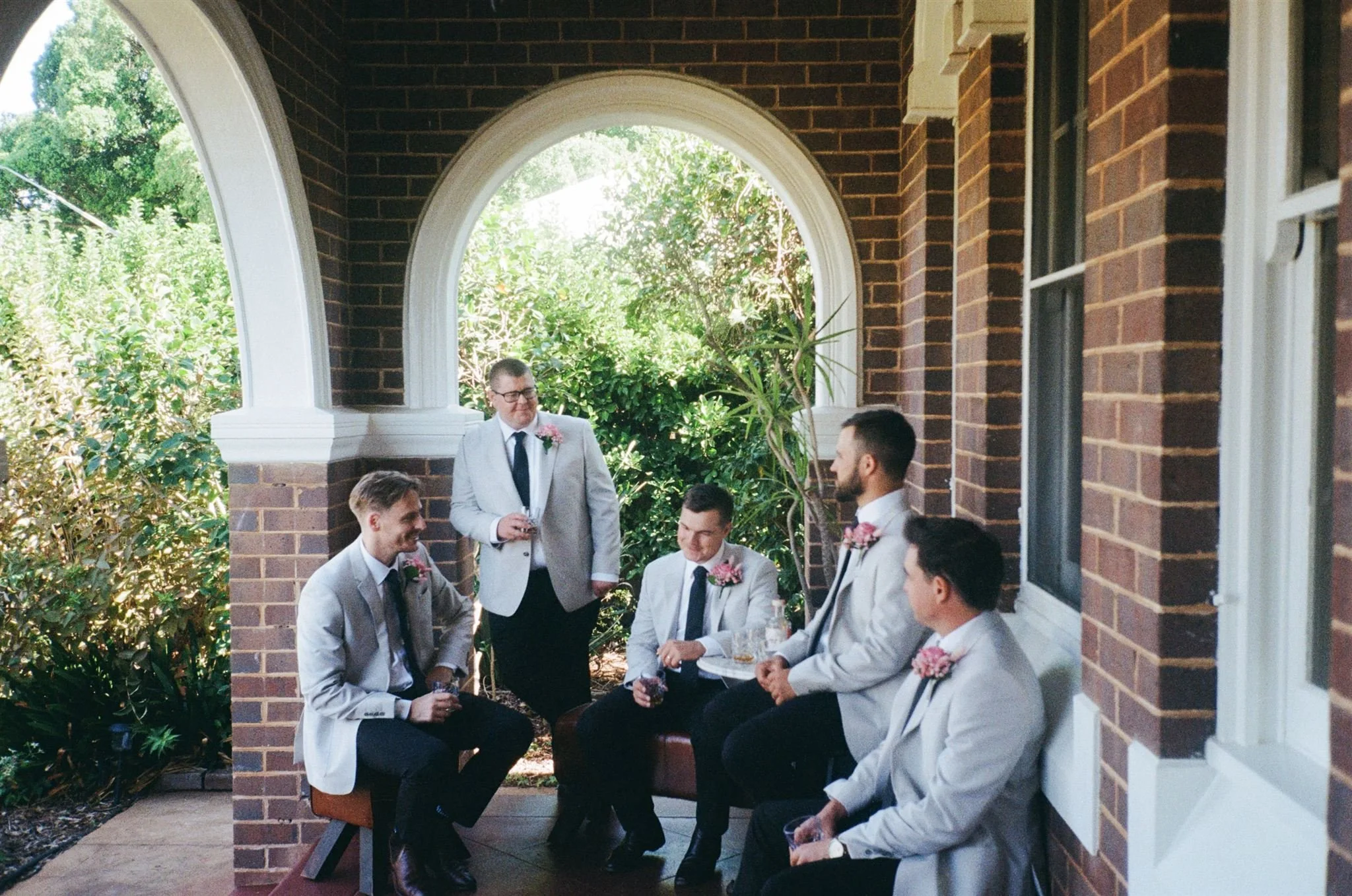 Six men in light gray suits with pink boutonnières, some sitting and some standing, having a conversation on a porch with brick columns and an arched opening, surrounded by greenery.