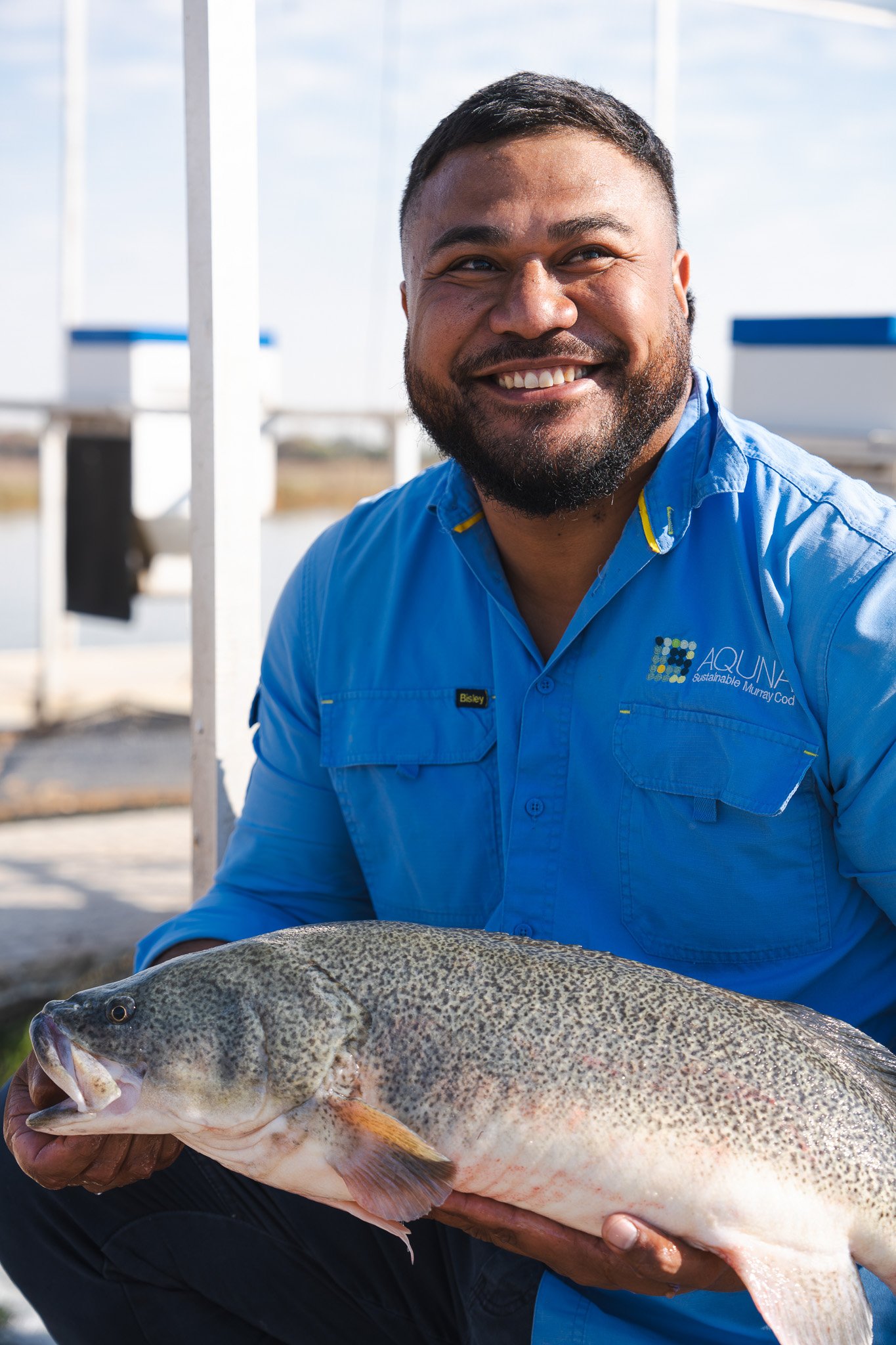A smiling man in a blue shirt holding a large fish outdoors near a water body.