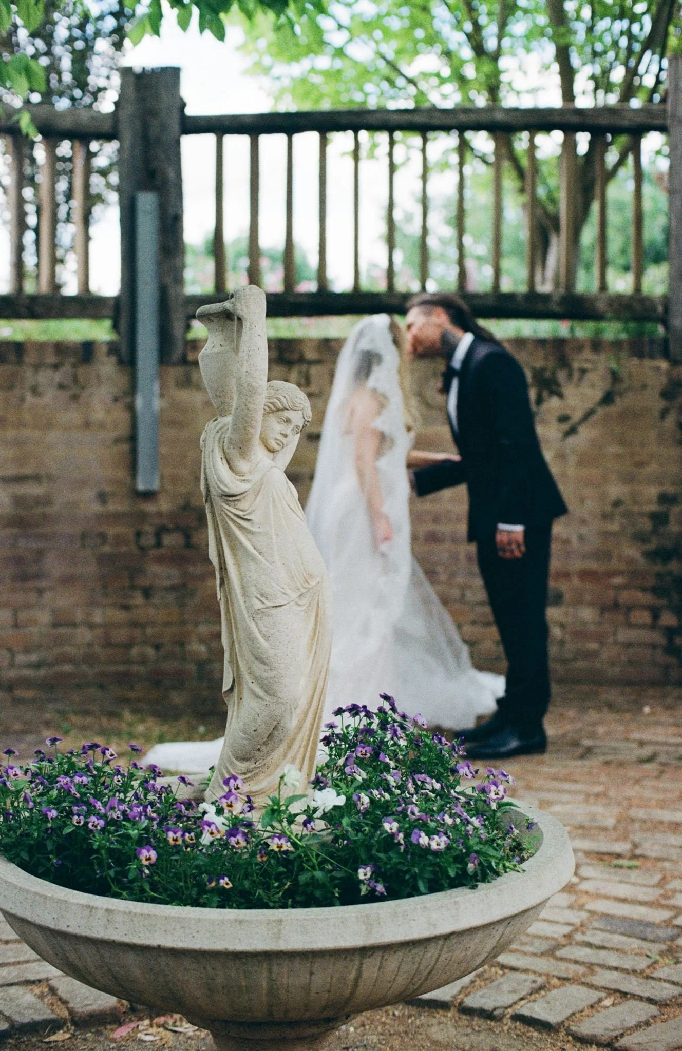 A wedding scene with a statue and bride and groom in the background. The statue is a classical sculpture of a woman holding an object, surrounded by purple and white flowers in a large planter. The bride in a white dress and veil, and the groom in a 