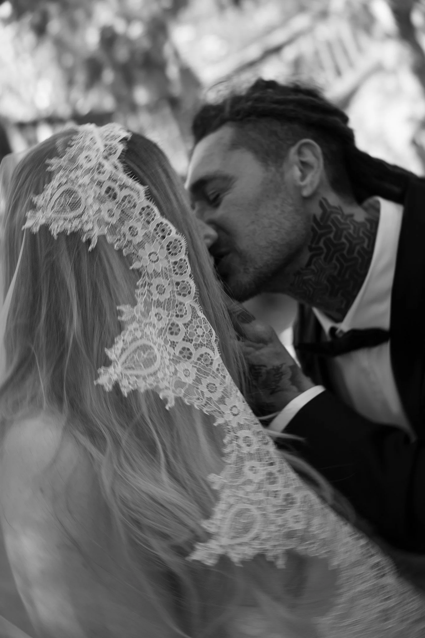 A black and white photo of a couple kissing, with the woman wearing a lace veil and the man dressed in a suit with tattoos visible on his neck.