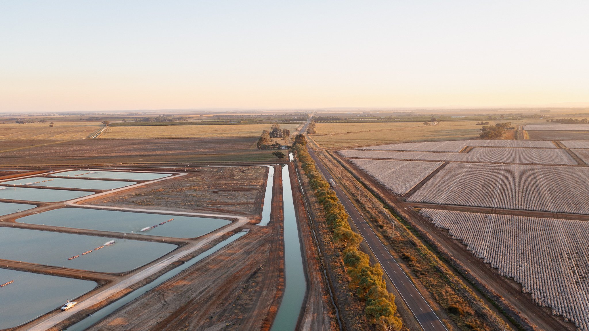 Aerial view of a landscape with fields, water canals, and a road with trees along the side, taken during sunset or sunrise.