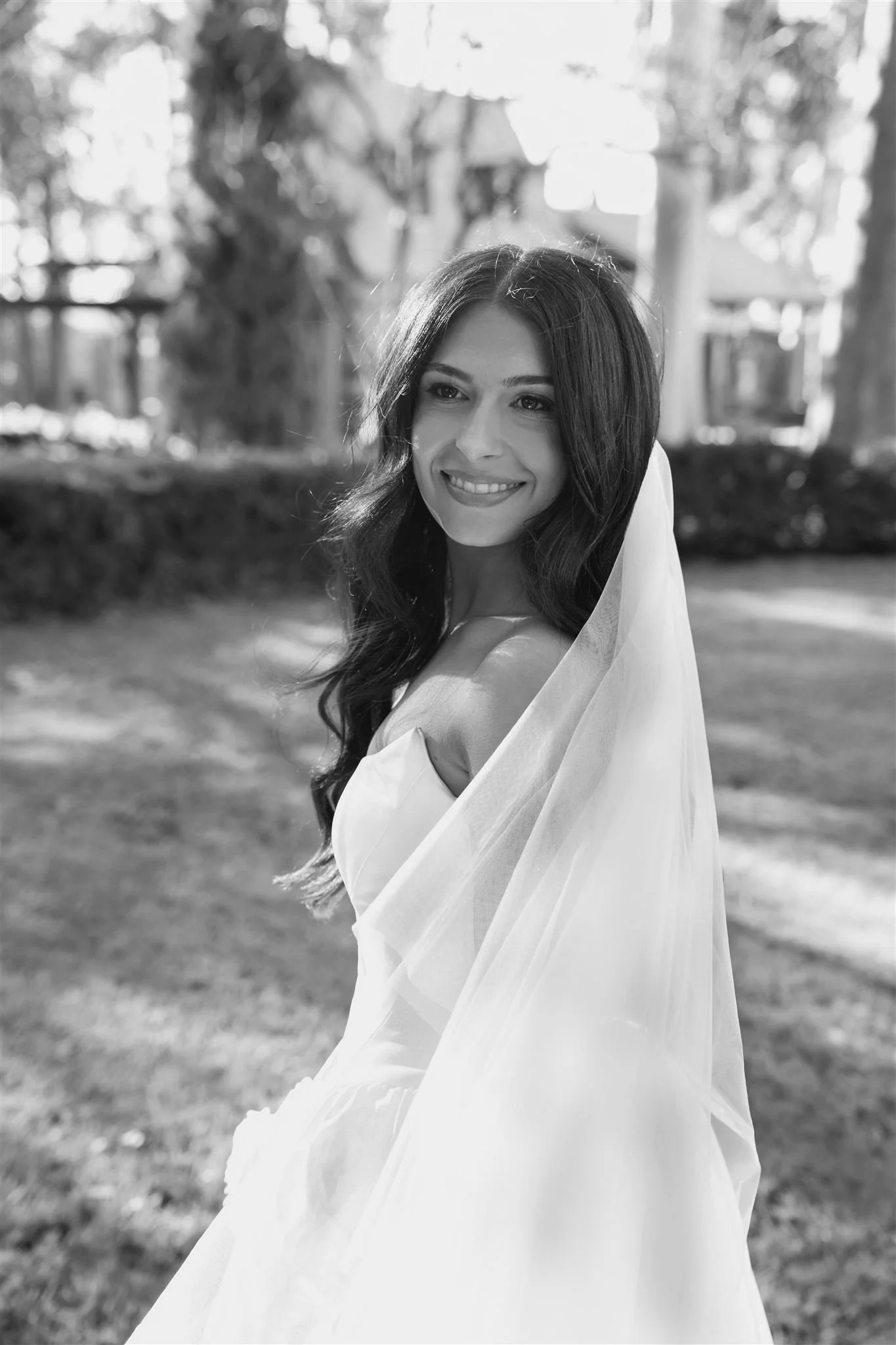 bride portrait, Black and white photo of a smiling woman outdoors, wearing a wedding dress and veil, with trees and buildings in the background.