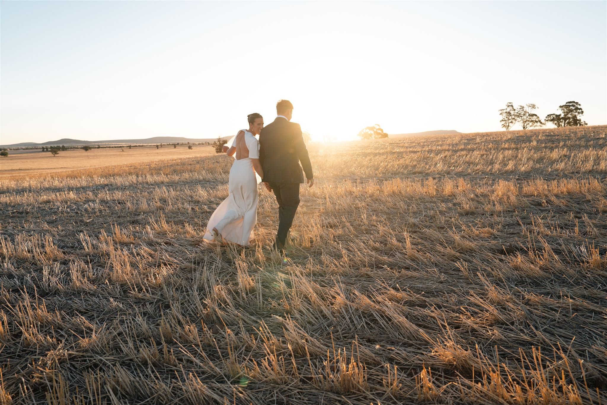 A couple walking hand in hand through a harvested wheat field at sunset, with a clear sky and distant trees.