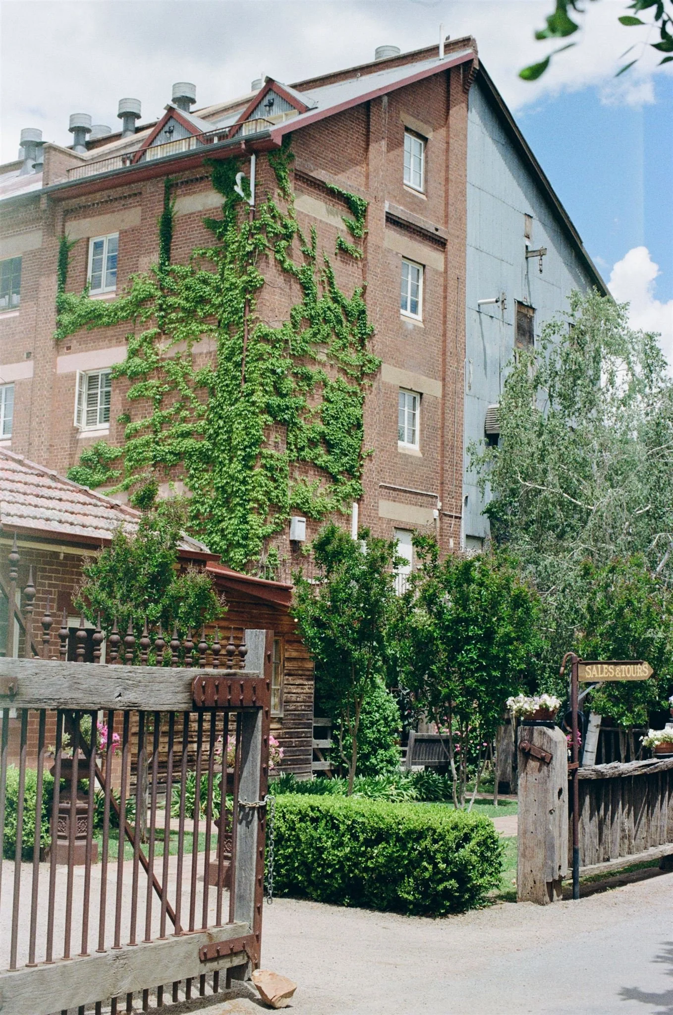 Junee Chocolate Licorice Factory, wedding. A multi-story brick building partially covered with climbing ivy, with a small yard enclosed by a rustic wooden fence, trees, and a sign that reads 'Sales & Tours'