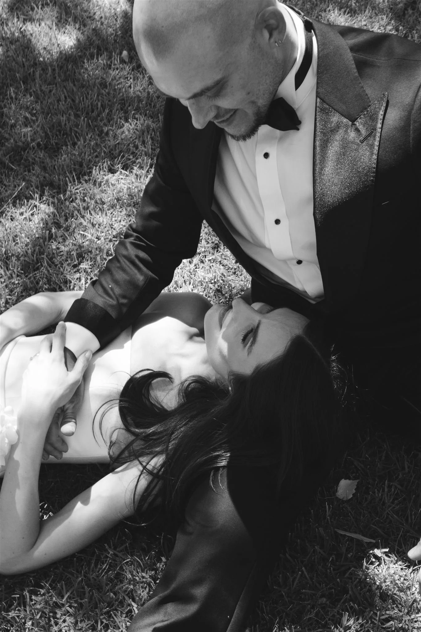 Griffith wedding photographer. Riverina photographer. A man in a tuxedo with a bow tie leaning over a woman in a sleeveless dress lying on the grass, holding hands.