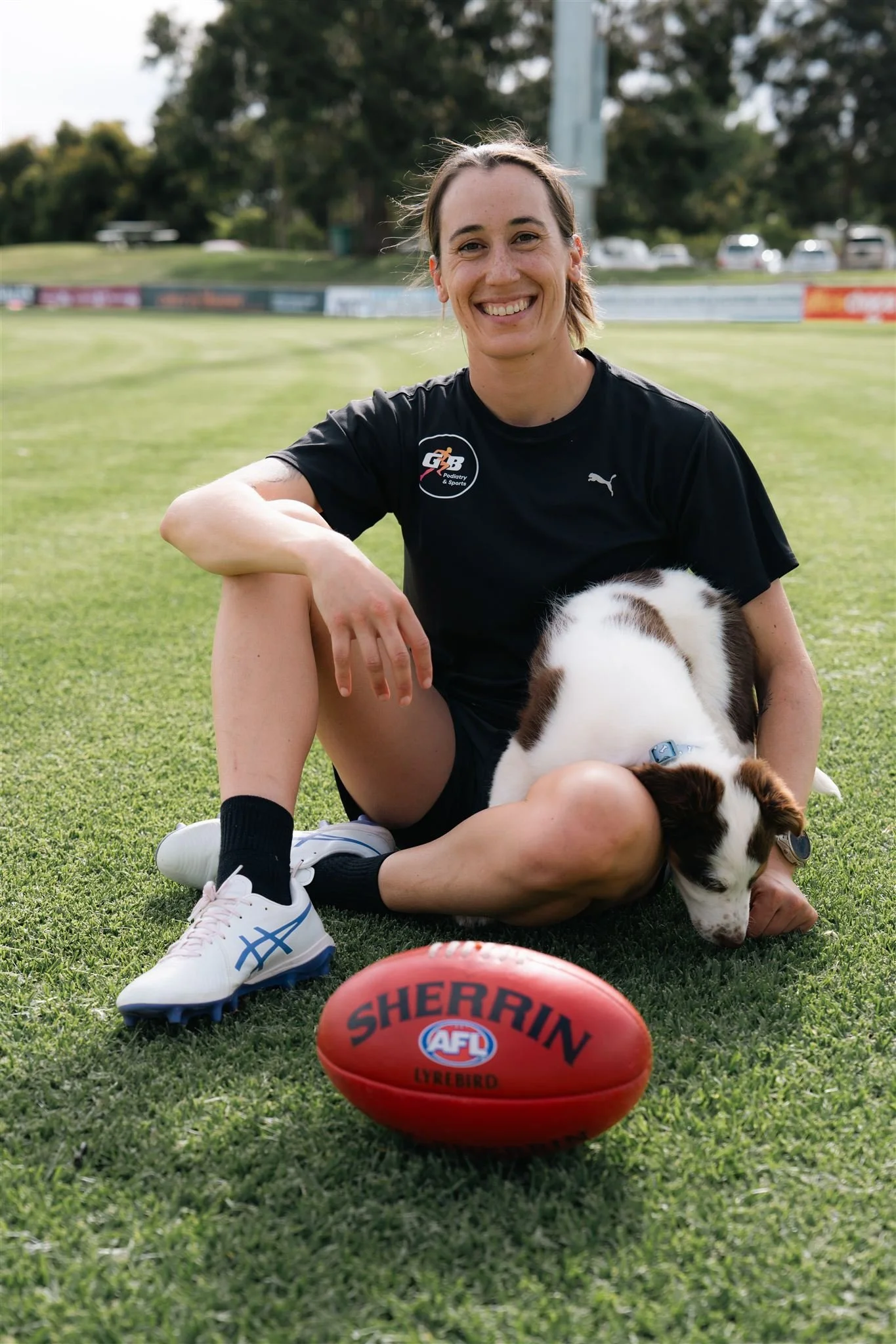 A smiling woman in sportswear sitting on a grassy field with a brown and white puppy, and an Australian rules football with the words 'Sherin' and 'AFL' on it.