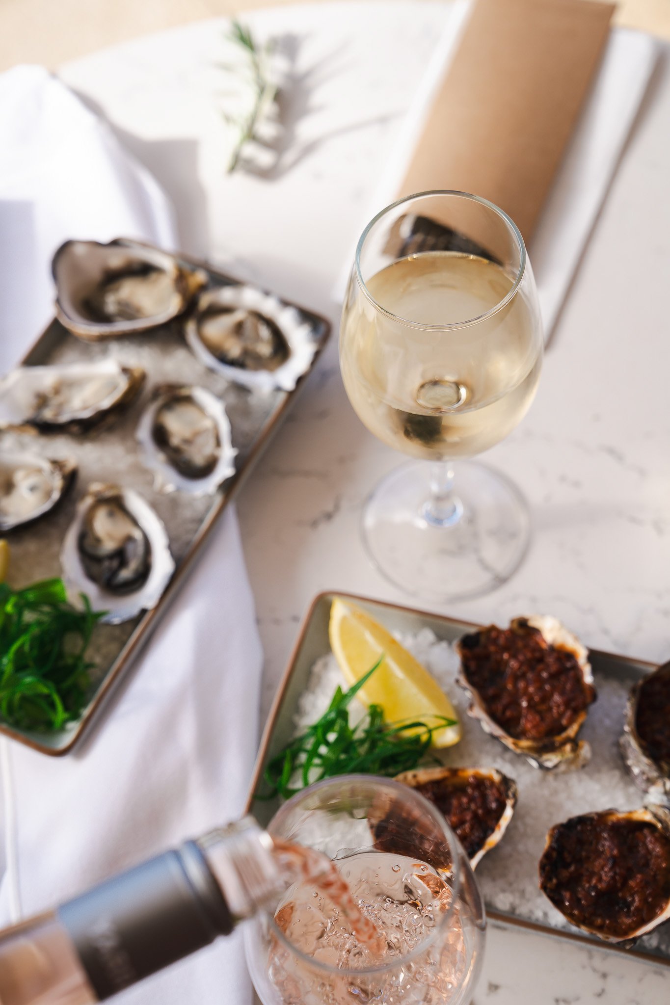 A glass of white wine, a glass of rosé, oysters on a tray, and a plate with lemon wedges and baked oysters on ice.