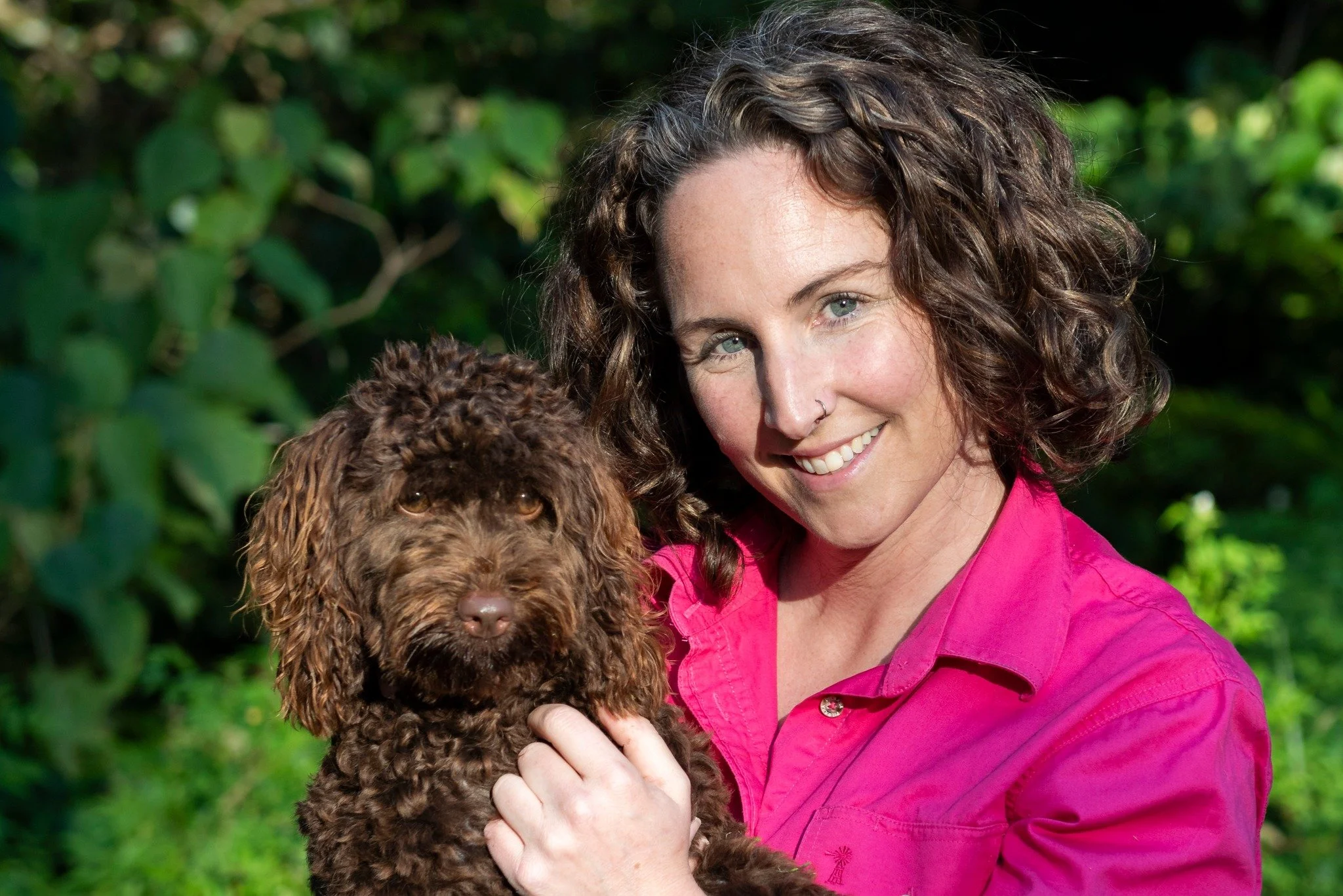 A woman with curly hair smiling and holding a brown curly-haired puppy outdoors.