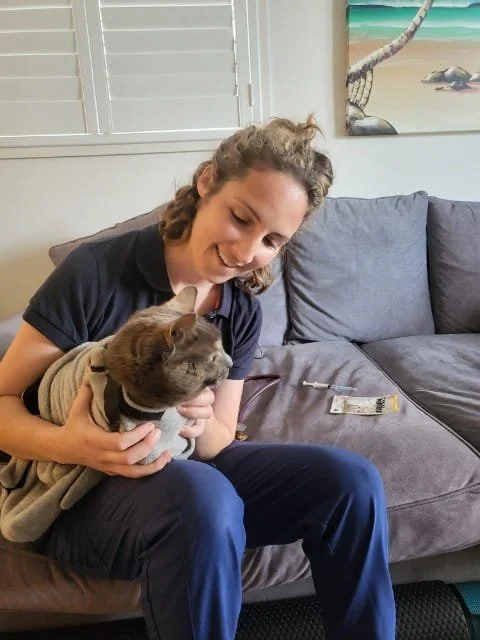 A young woman sitting on a couch holds a gray cat wearing a beige hoodie, smiling and looking at the cat inside a cozy living room.