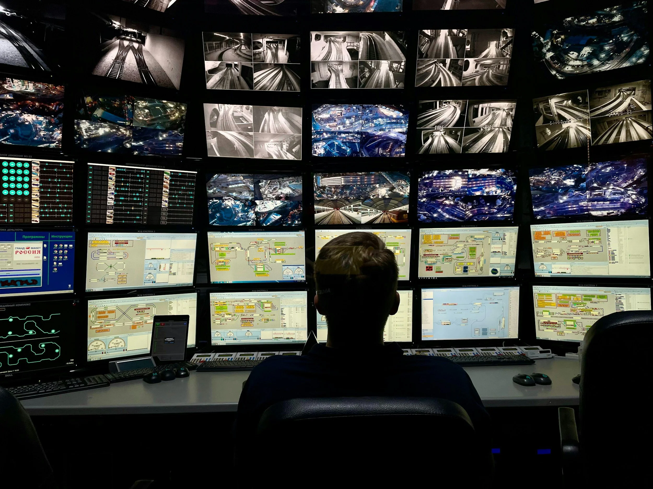 A control room with dozens of monitors displaying train tracks, tunnels, and cityscapes, with individuals monitoring the screens.
