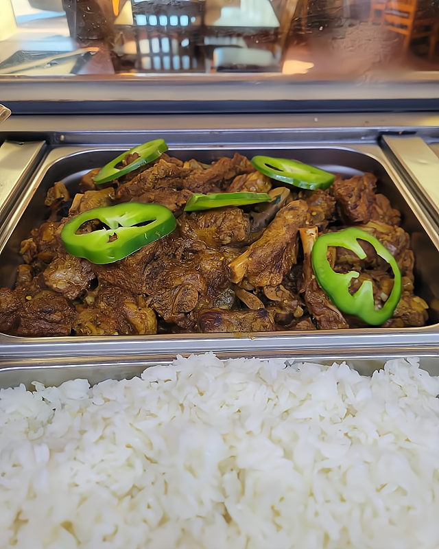 Cooked beef ribs topped with sliced green bell peppers in a buffet serving tray, with white rice in a separate tray in front.