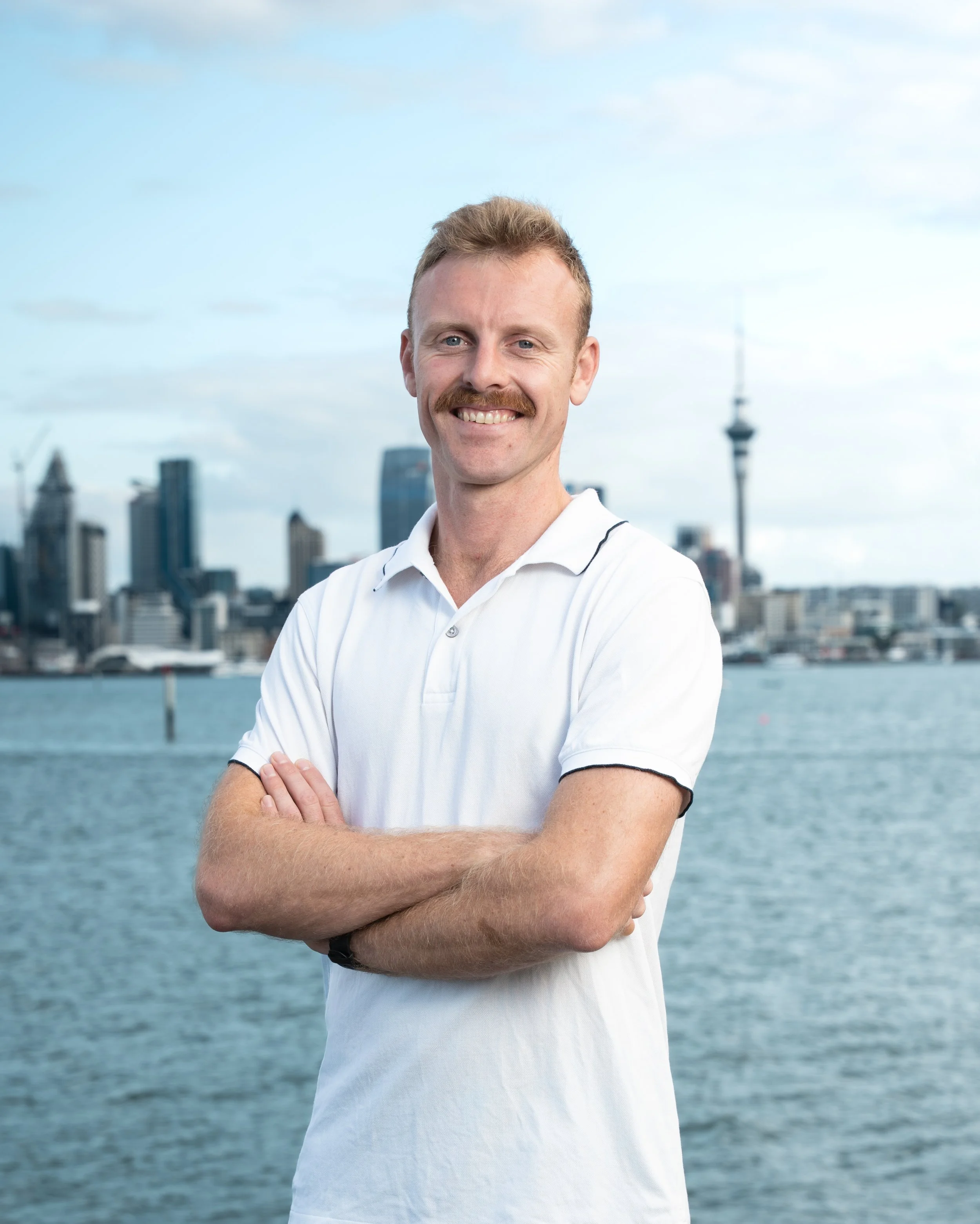 A man with red hair, a mustache, and blue eyes smiling with his arms crossed, standing outdoors near water with a city skyline and a tower in the background.
