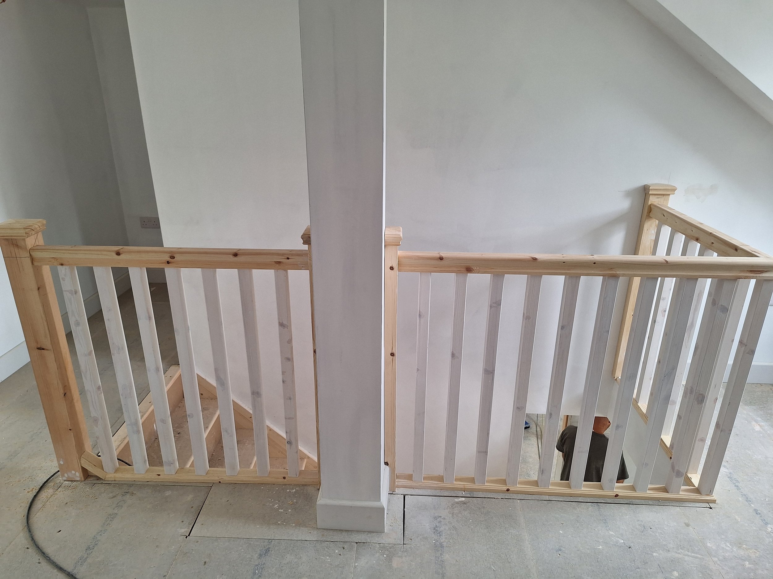 Interior of a house under construction featuring a small wooden staircase with a railing, dividing two areas, with a partial view of a worker at the bottom.