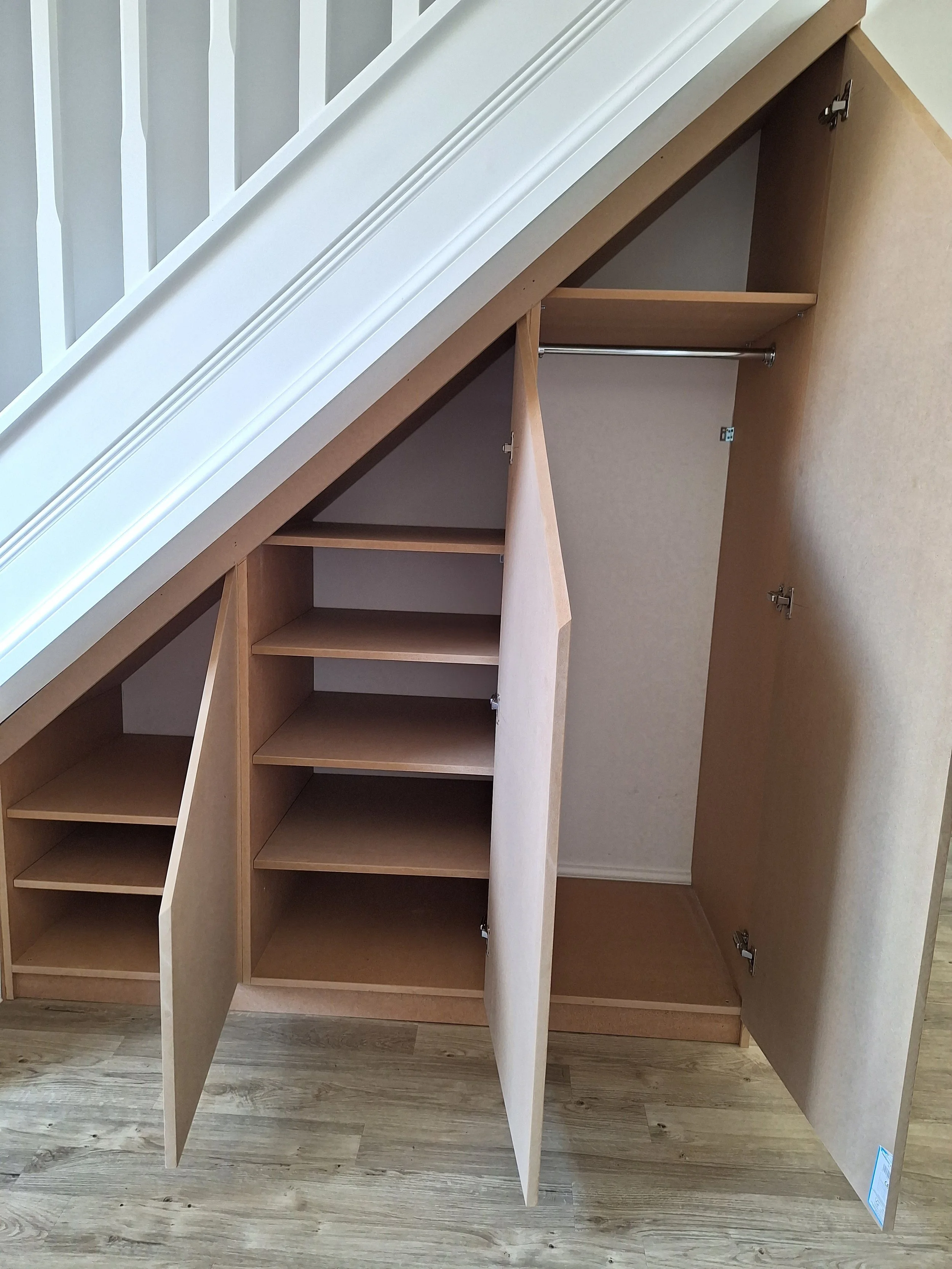 Open built-in under-stair storage with three sections: a small shelving unit on the left, a middle section with shelves behind a door, and a right section with a hanging rod and an open space beneath.