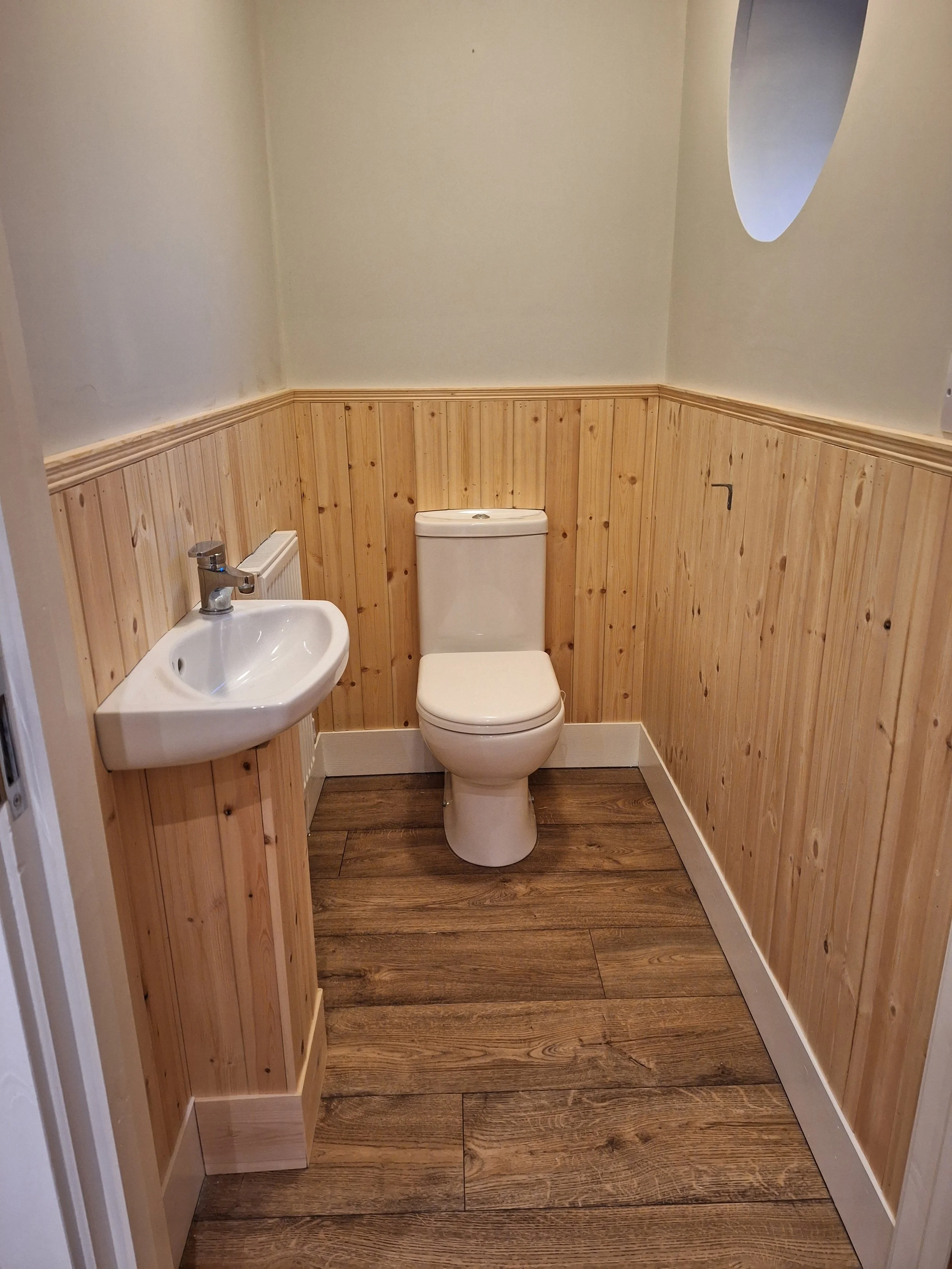 Small bathroom with wood paneling on the lower half of the walls, a white toilet, a small white corner sink with a silver faucet, and a small oval window near the ceiling.