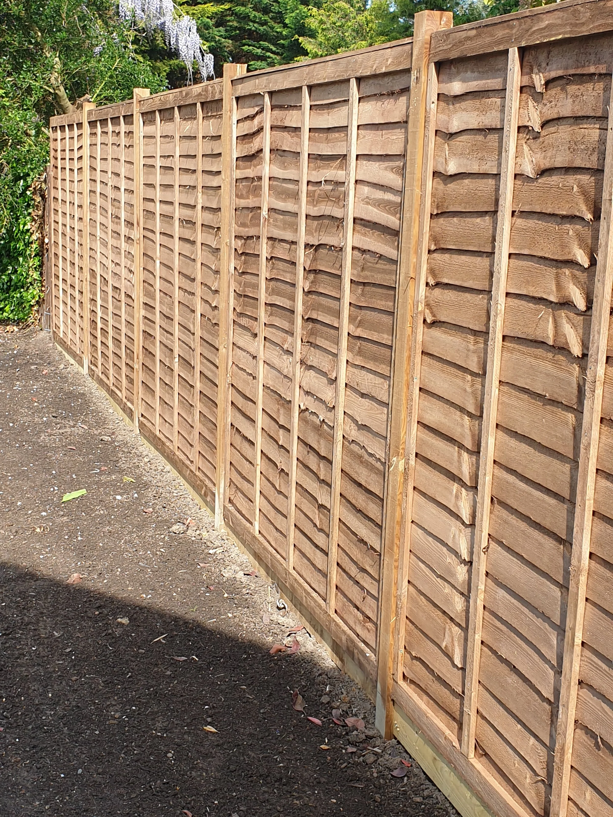 A newly constructed wooden fence with vertical and horizontal slats, installed along a dirt pathway in a garden setting, with trees and plants in the background.
