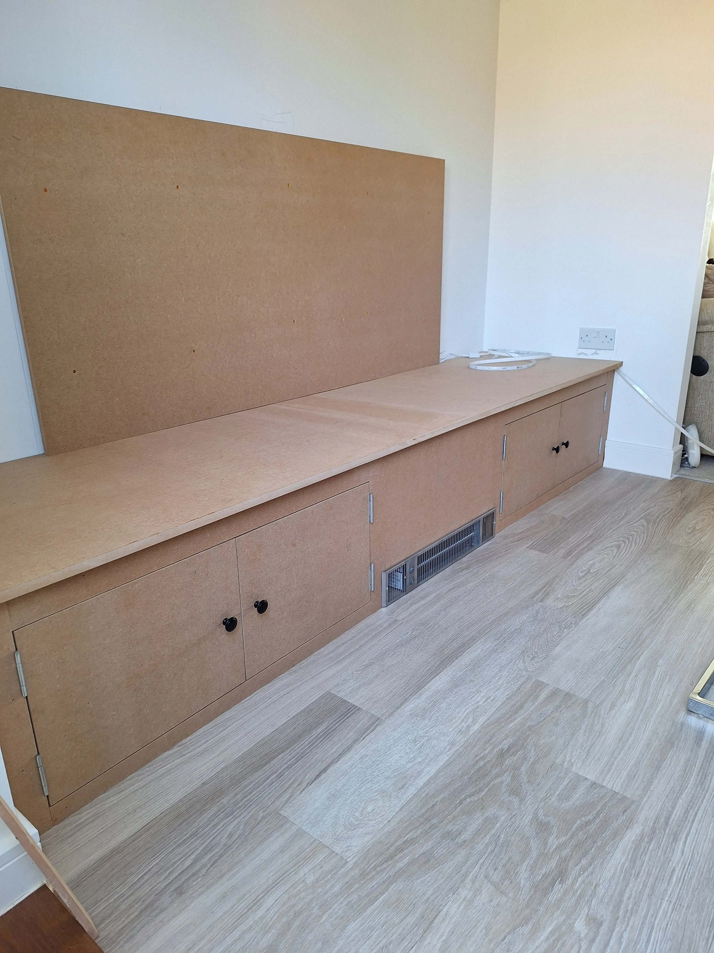 Unfinished wooden built-in cabinet with cabinet doors and a large corkboard on top, located in a room with light-colored wood flooring and white walls.