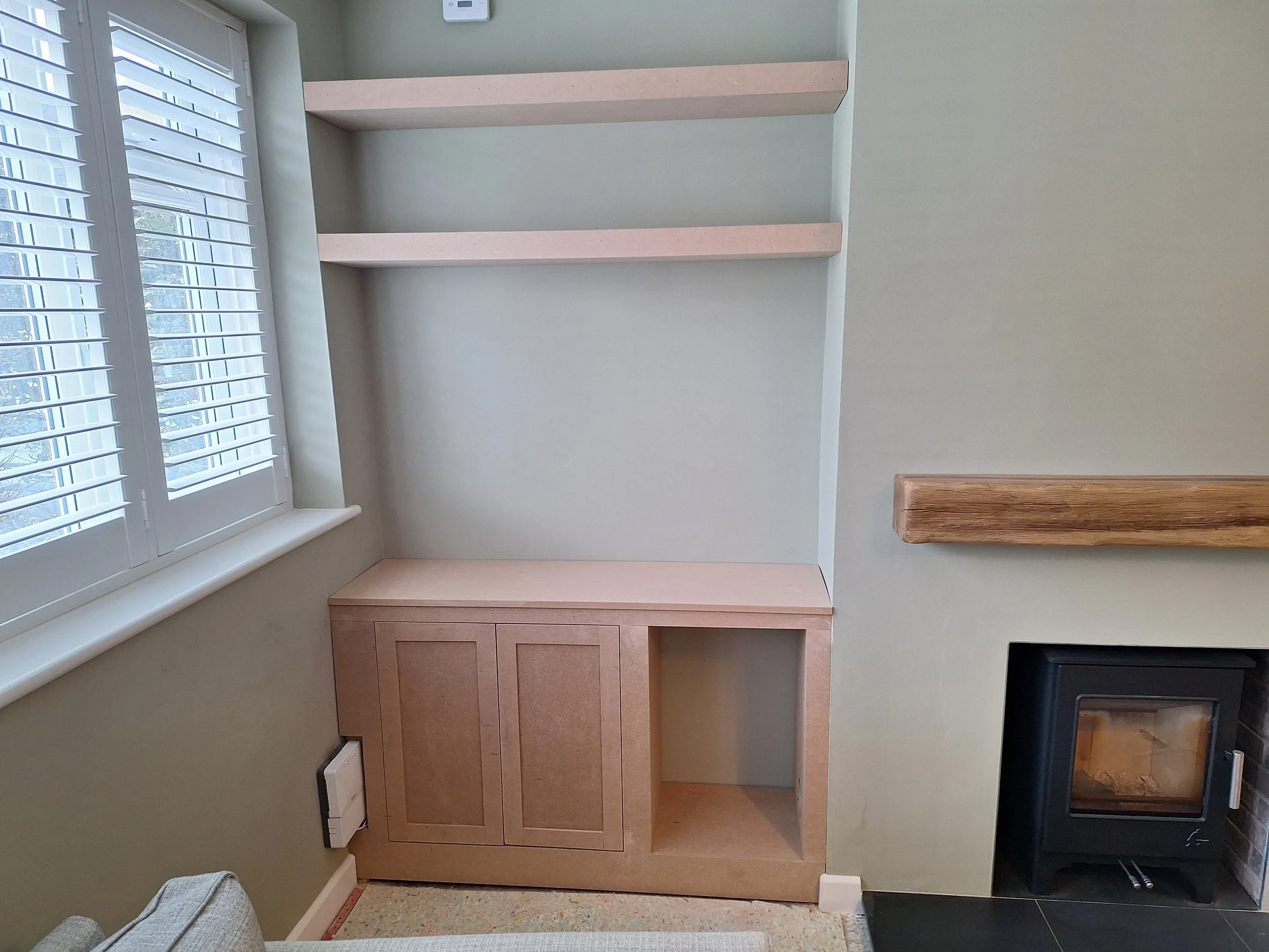 Empty beige wooden cabinetry next to a window with white blinds and a fireplace with a wooden mantel in a living room.