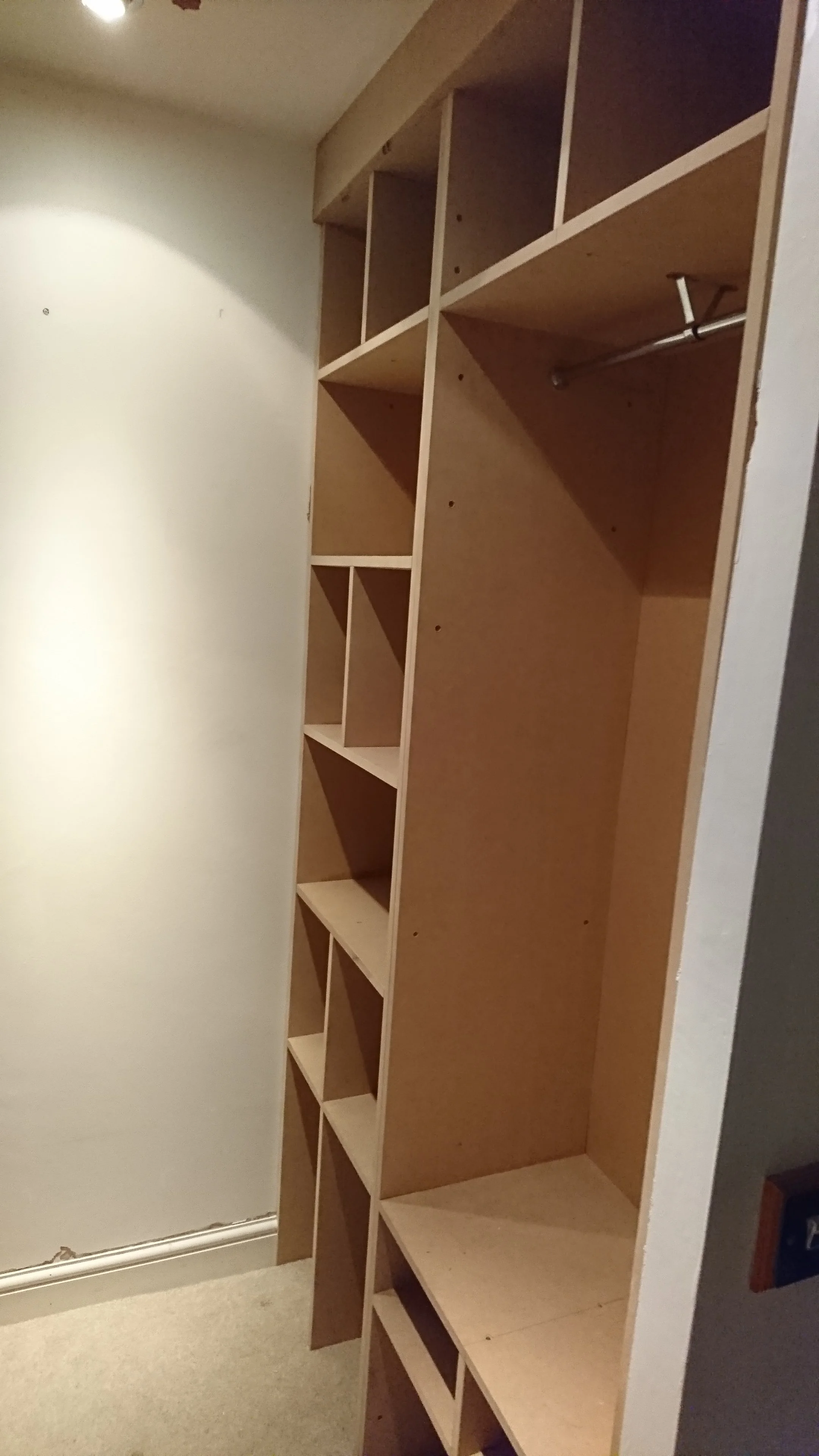 Unfinished wooden built-in closet with multiple compartments and a hanging rod, situated in a room with white walls and carpeted floor.