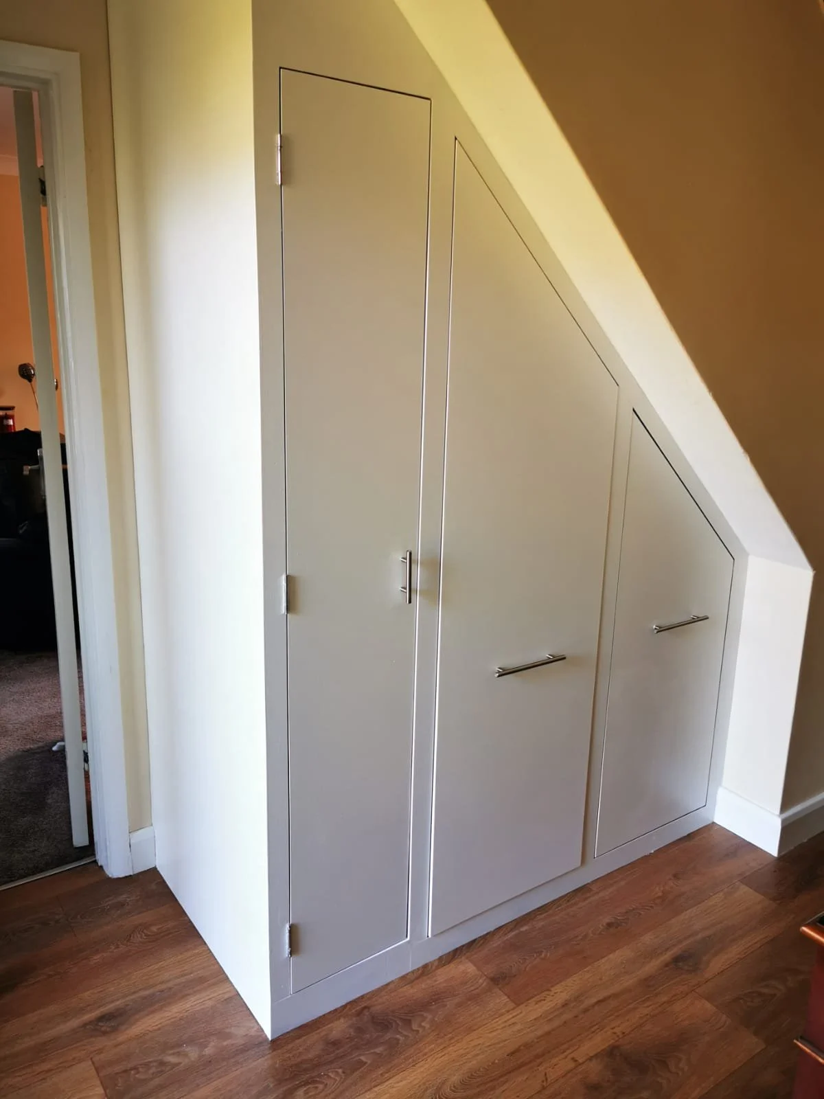 Corner cabinet with slanted doors in an interior space, adjacent to a hallway with wooden flooring.