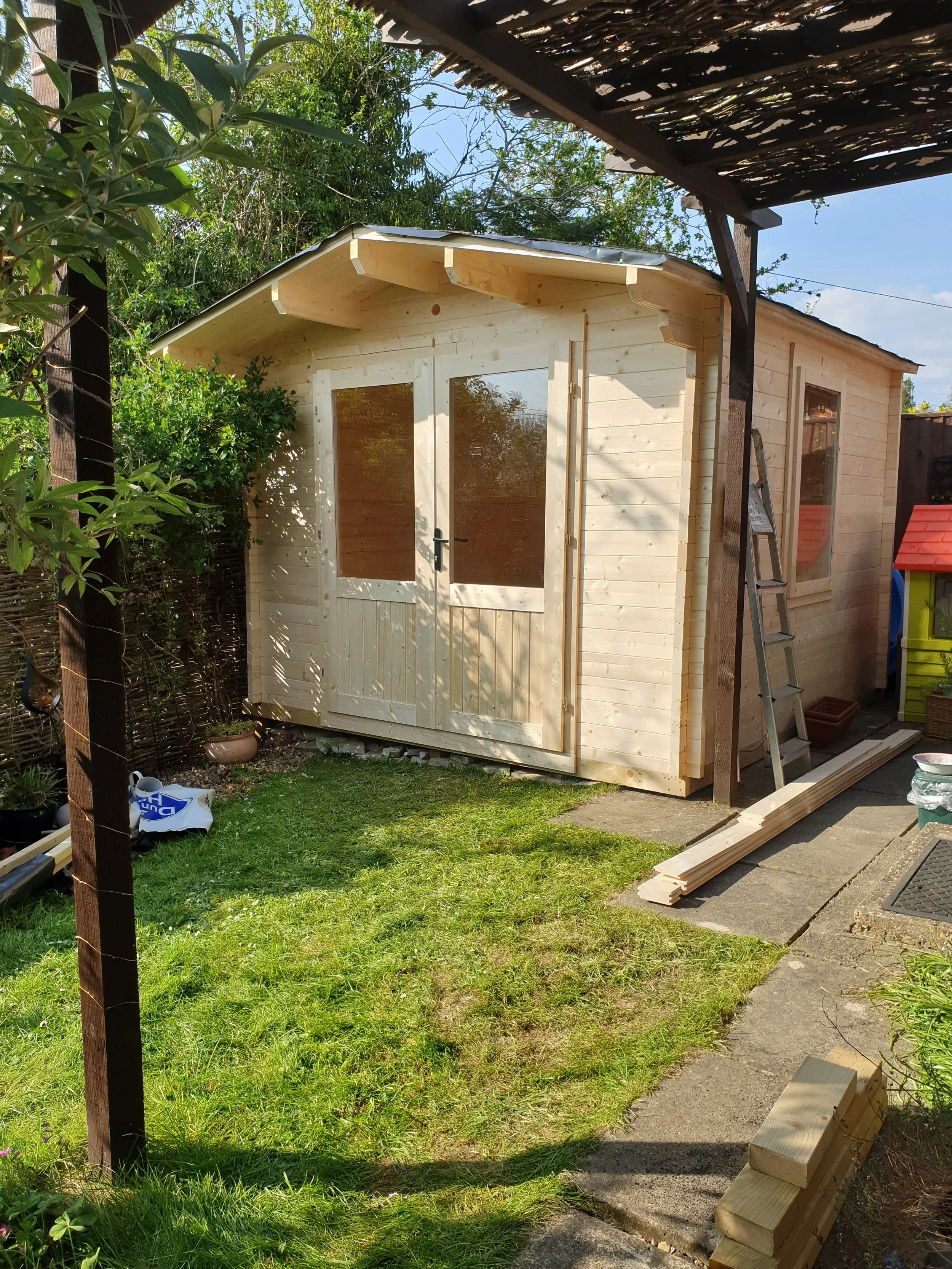A small wooden garden shed with a double door and a window, situated on a grassy lawn in a backyard with plants, trees, and a wooden fence. Construction materials and tools are nearby, indicating the shed is unfinished or being built.
