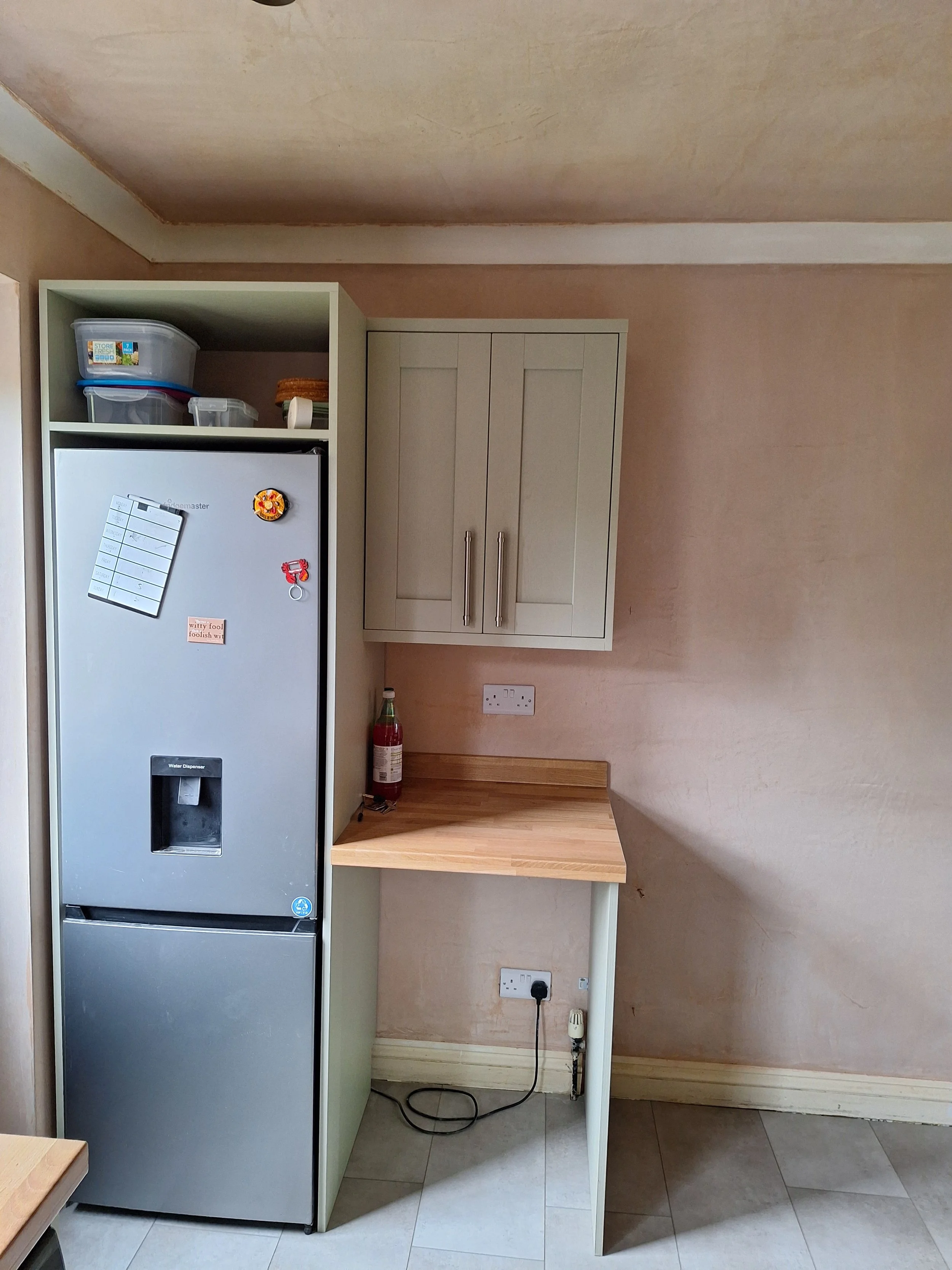 A kitchen corner with a silver refrigerator, white cabinets, and a small wooden countertop, with an electrical outlet on the wall.