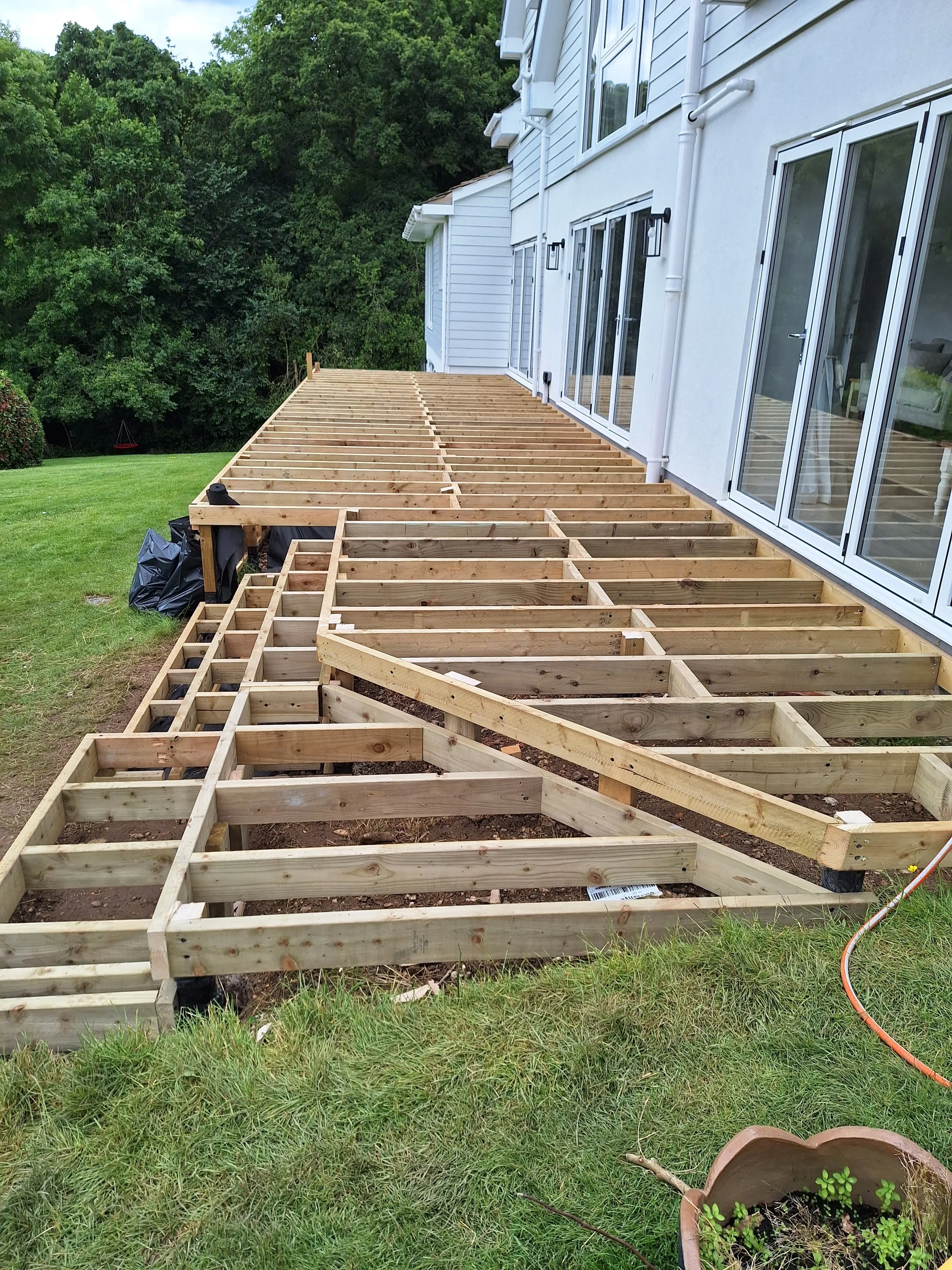 Wooden frame construction of a deck attached to a white house, with sliding glass doors leading inside, and a green grassy yard with trees in the background.
