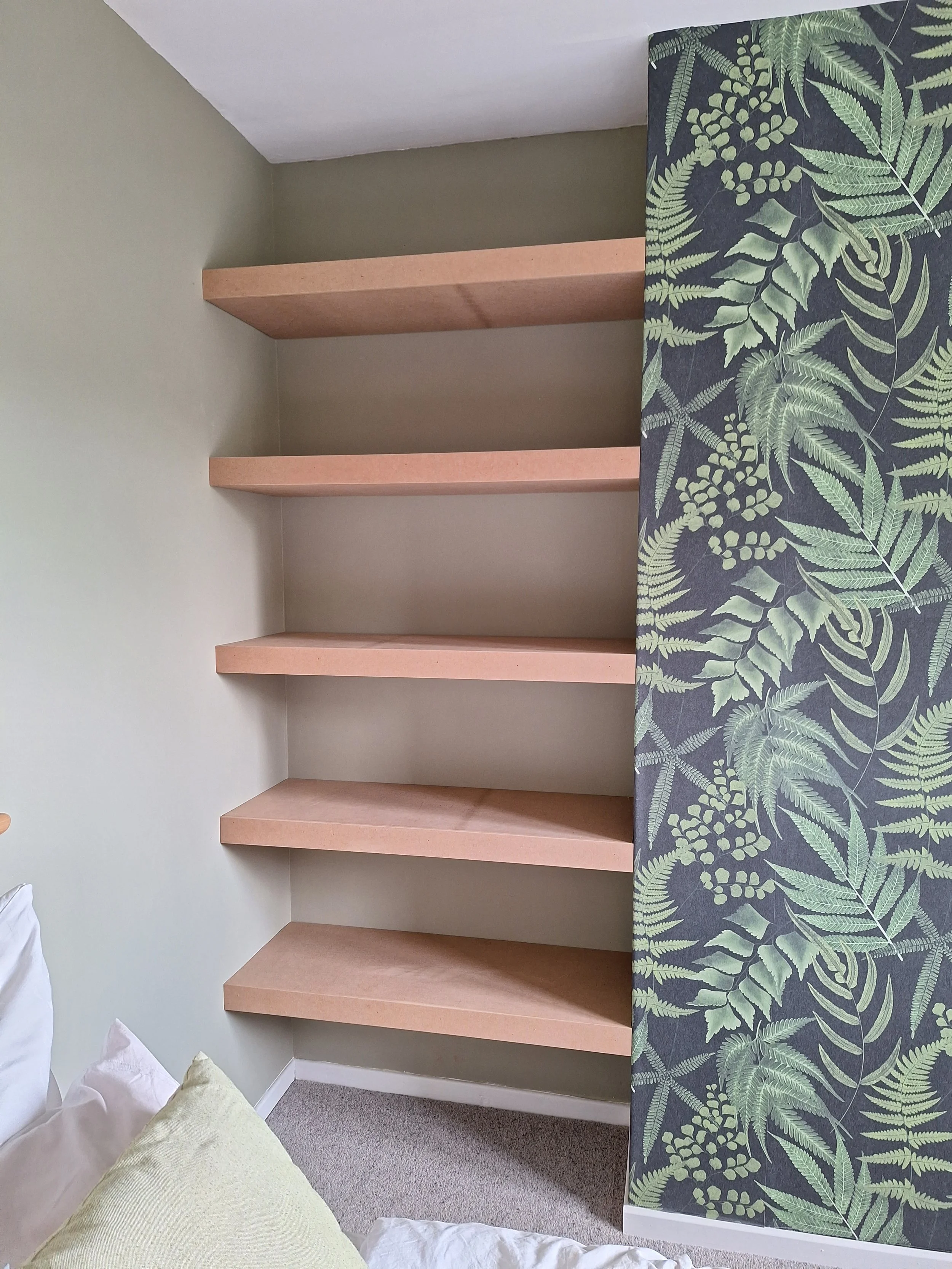 Empty set of pink floating shelves on a beige wall with a tropical leaf patterned wallpaper on the side.