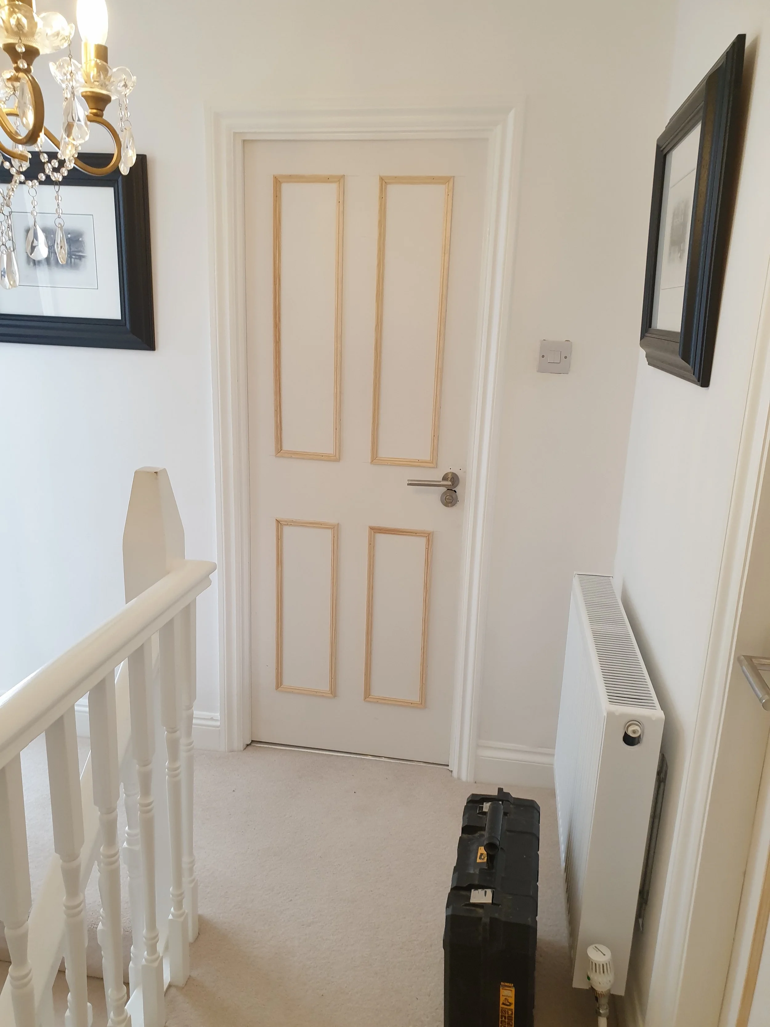 Hallway with closed door, framed pictures on wall, radiator, staircase railing, and a toolbox on beige carpet.