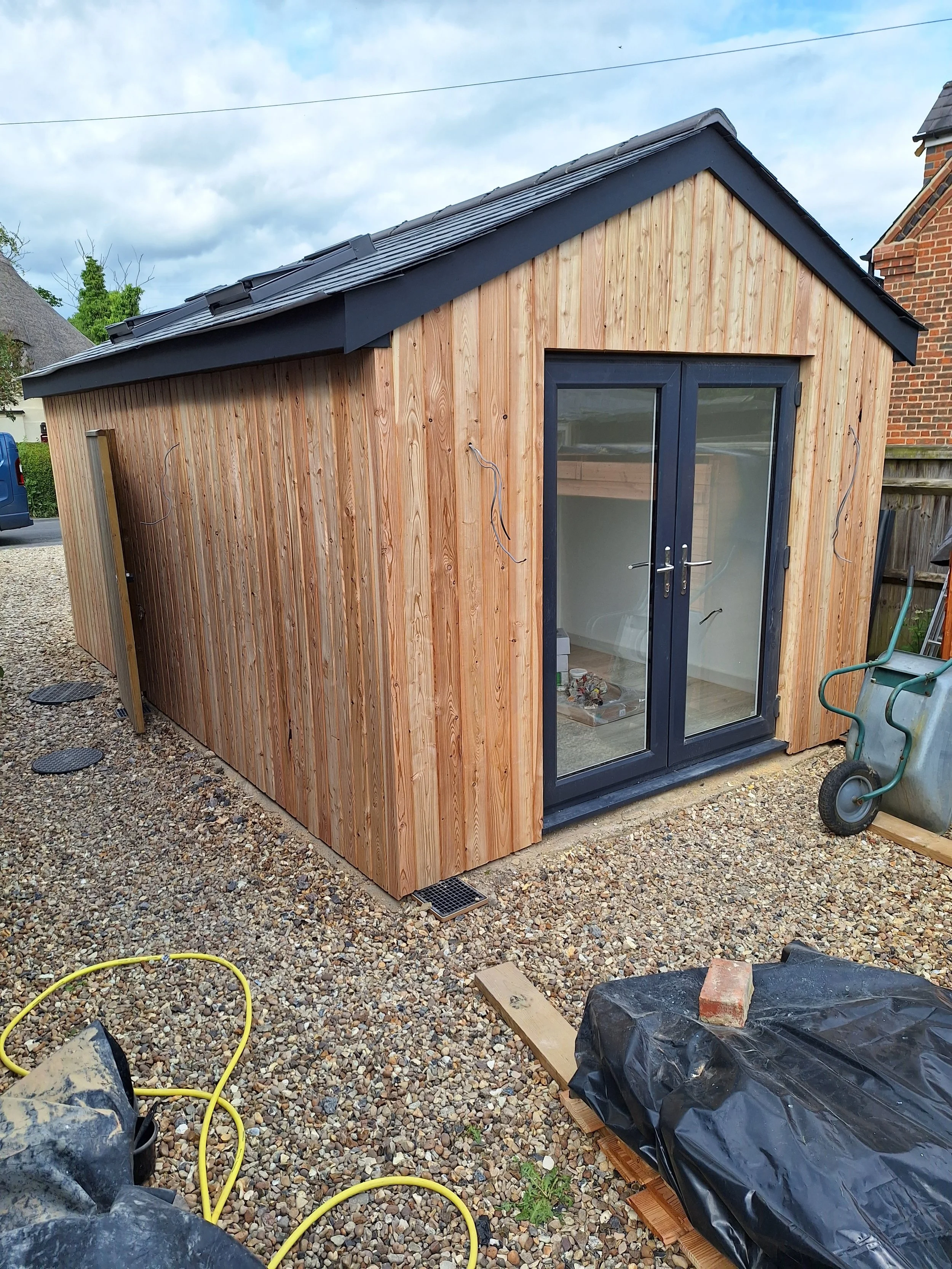 A small wooden shed with black-framed glass double doors, placed on a gravel surface, under construction with visible wiring and some supplies around.
