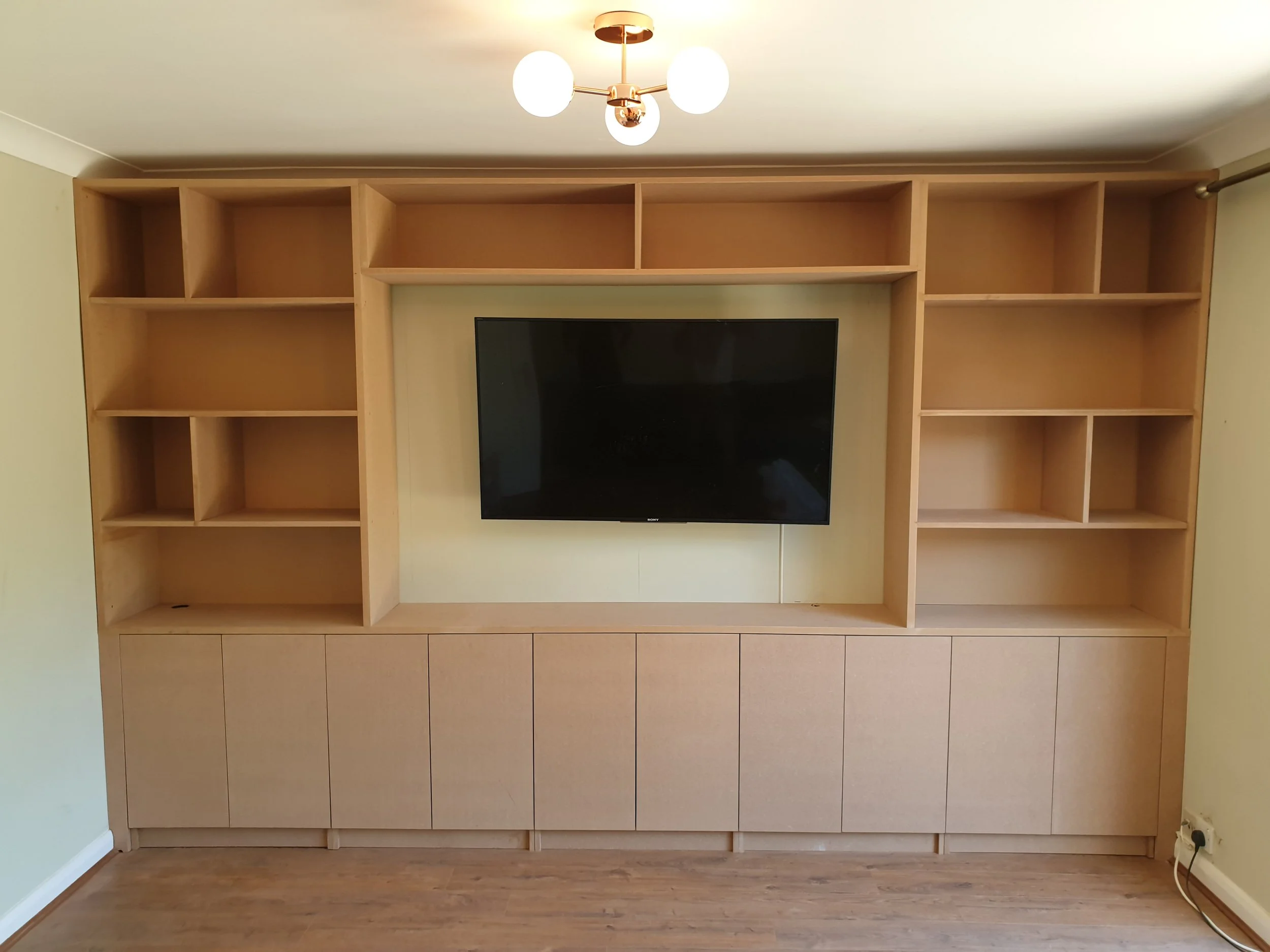Empty wooden entertainment center with a large flat-screen TV in the middle, open shelves above and closed cabinets below, in a room with beige walls and a wooden floor.