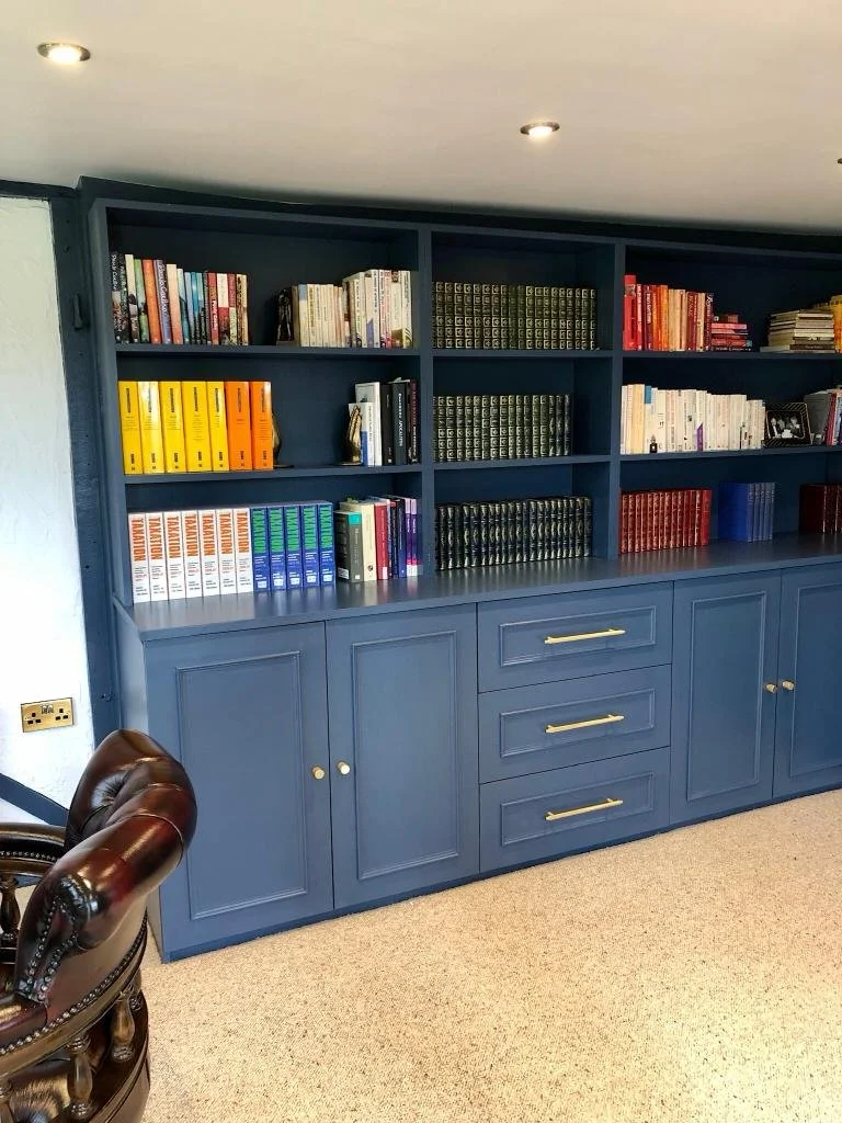 Blue built-in bookshelf filled with various books and decorative items, with a blue cabinet underneath and a brown leather armchair partially visible in the foreground.