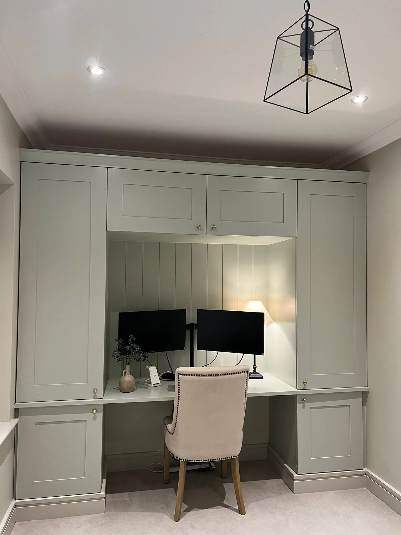 A home office with a built-in light green desk and cabinetry, two computer monitors, a beige upholstered chair, a small lamp, a vase with flowers, and a ceiling light fixture.