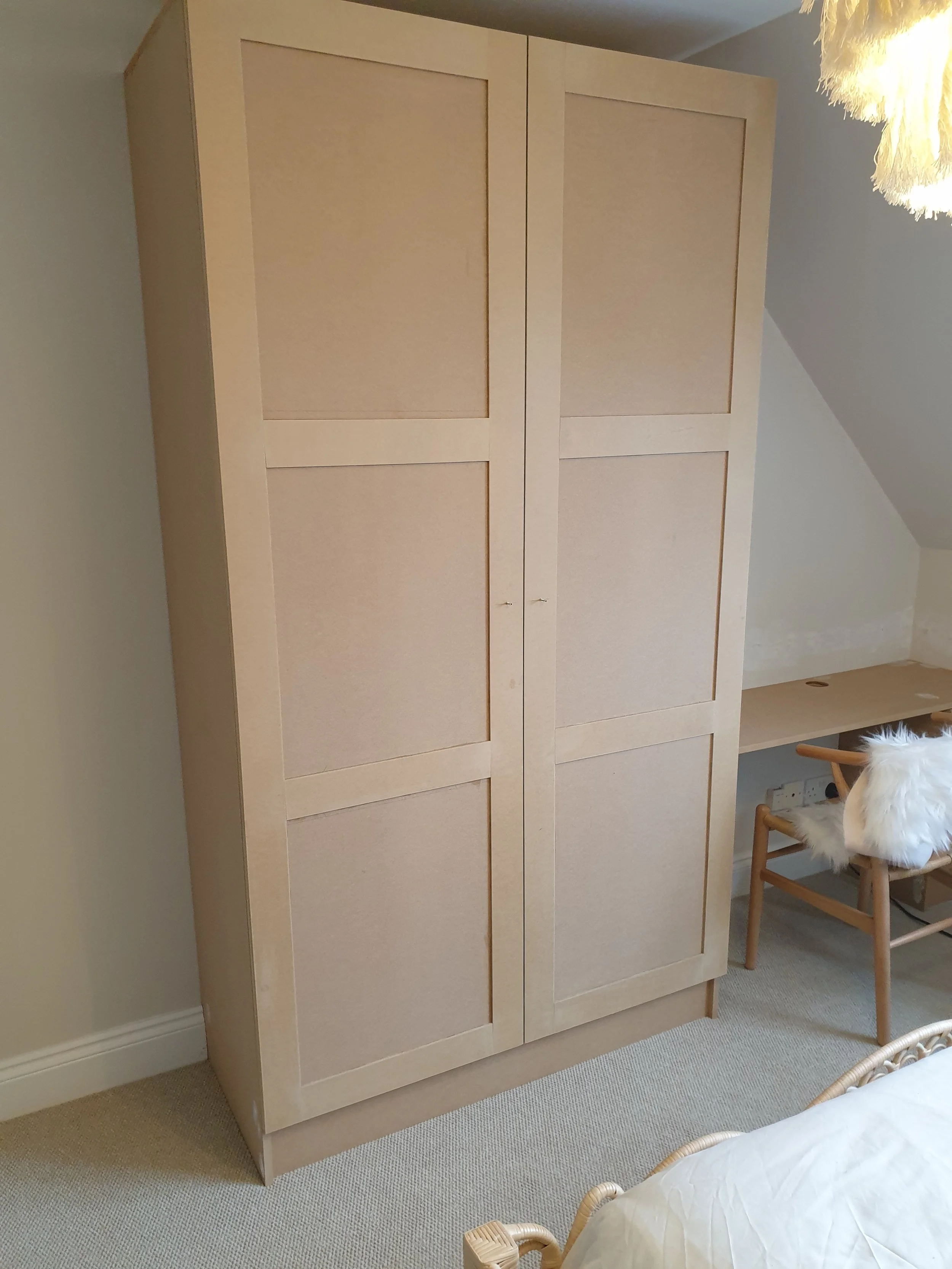 A large beige wooden wardrobe in a bedroom, next to a small desk and chair with a faux fur throw, part of a bed with a white duvet, and beige carpet.