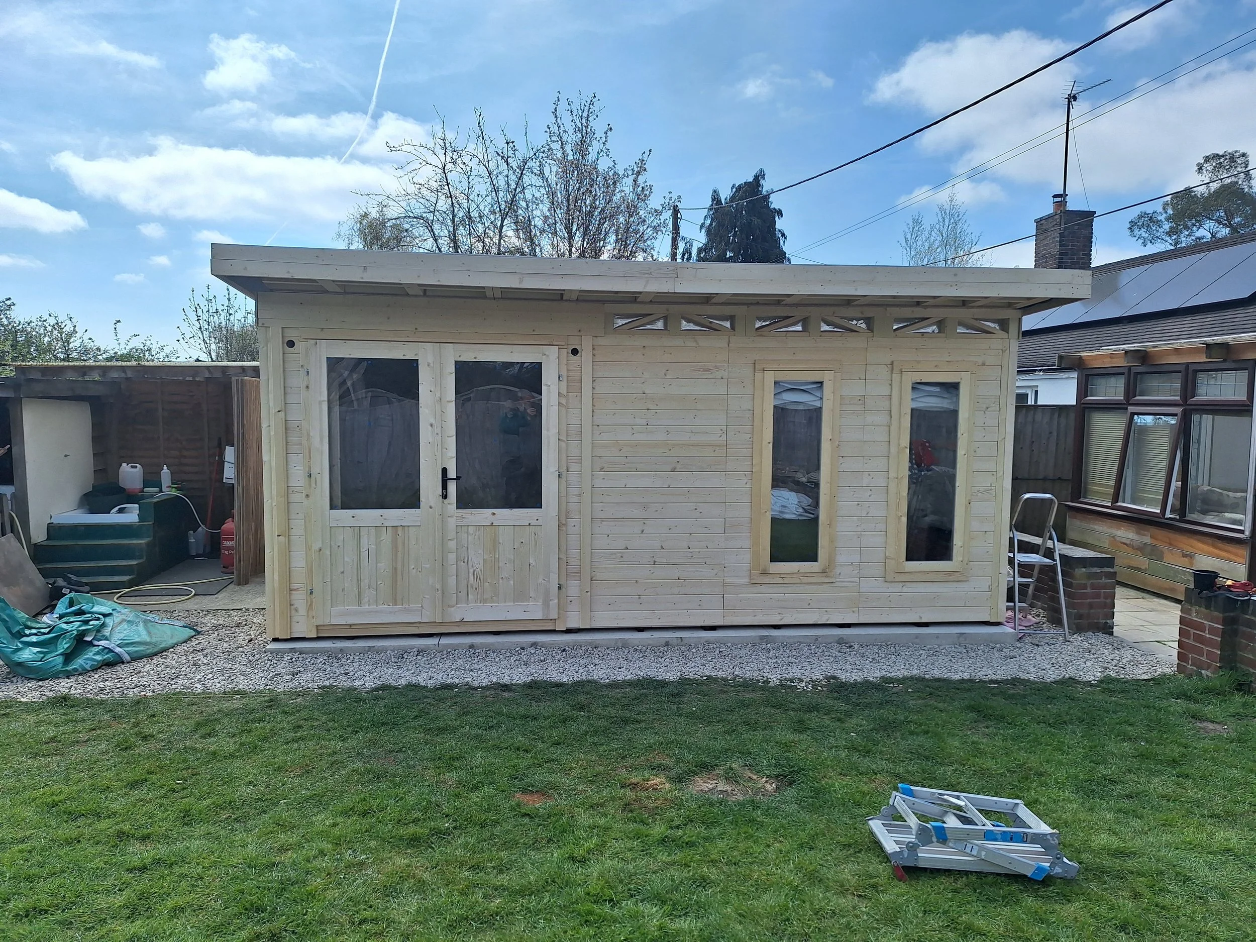 A small wooden shed under construction, with double doors and three narrow windows, set on a gravel base in a backyard with a grassy lawn.