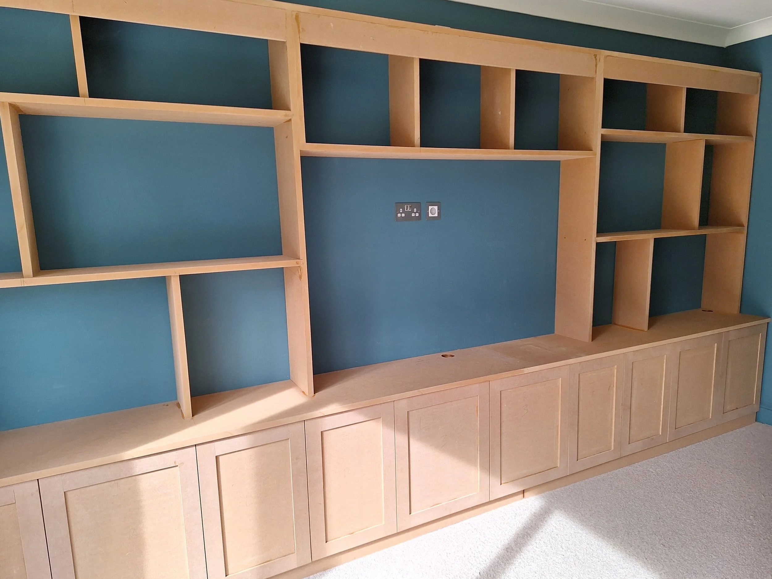Unfinished wooden built-in shelving unit mounted on a blue wall, with multiple open shelves and cabinets at the bottom.