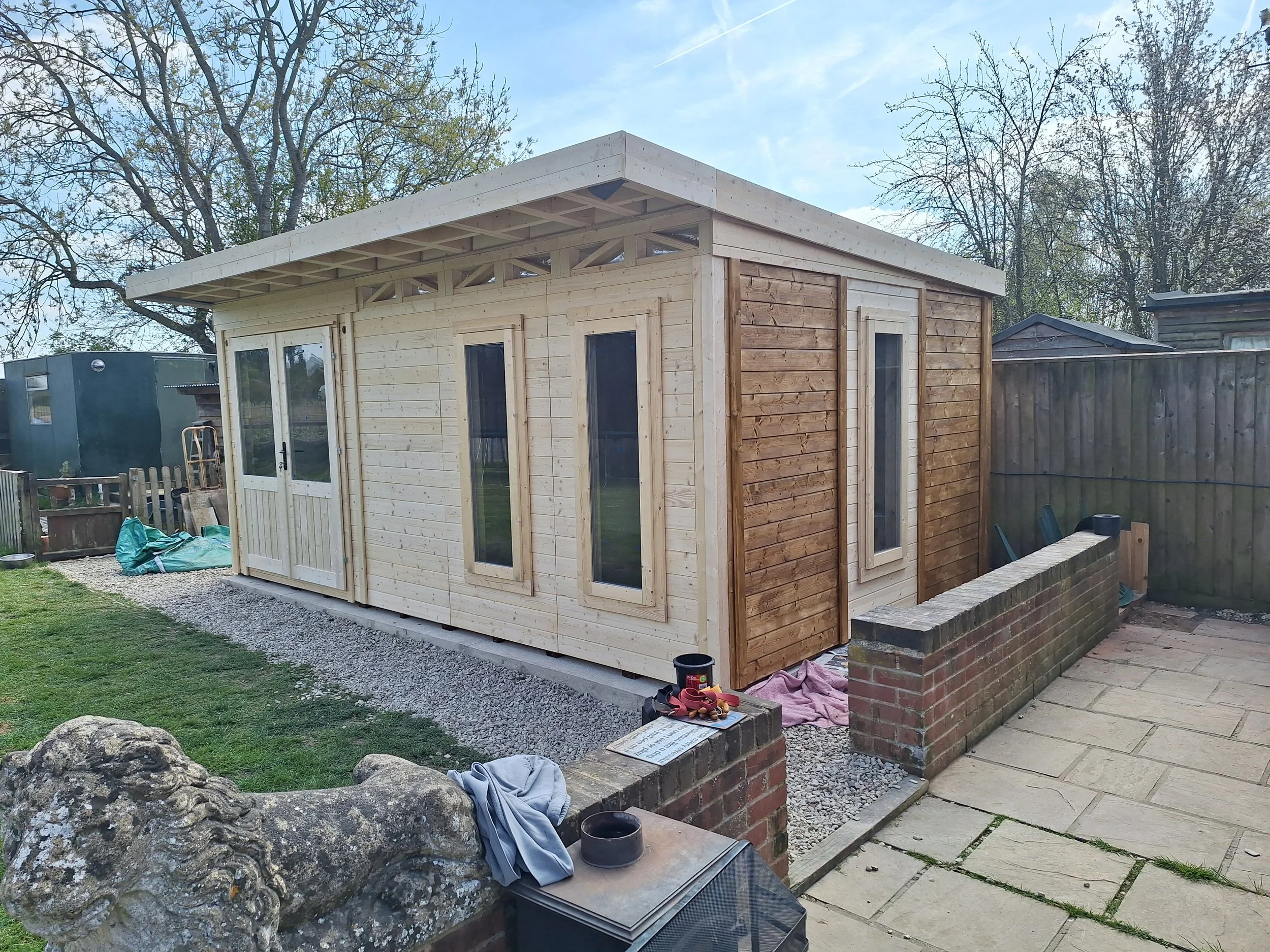 A small wooden shed under construction in a backyard, with some tools and materials nearby, surrounded by a garden with grass, gravel, and paved area, with trees and fence in the background.